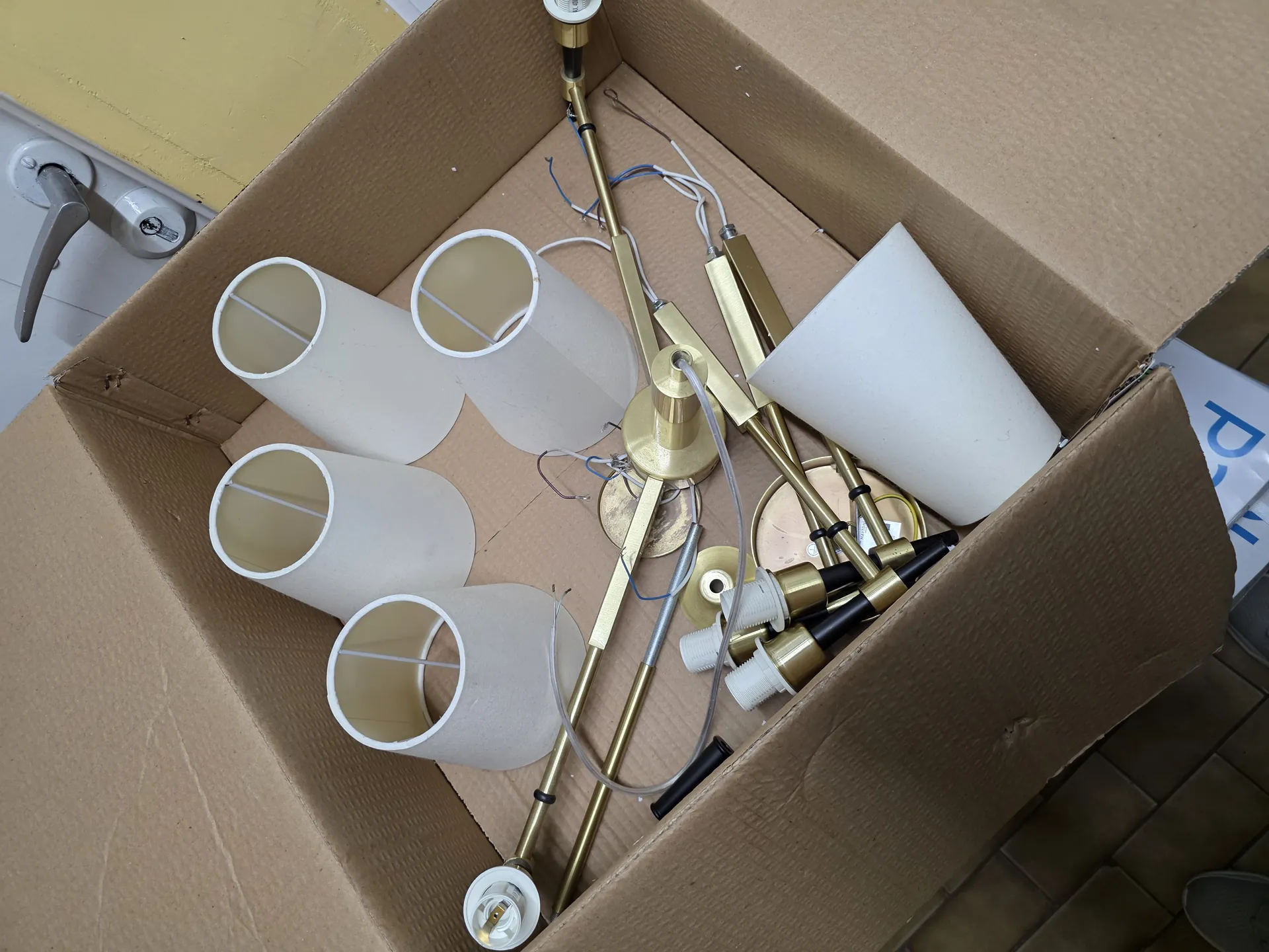 Otwarte kartonowe pudełko zawierające cztery beżowe cylindryczne abażury, mosiężne elementy konstrukcyjne lampy, przewody elektryczne (niebieskie i białe), białe plastikowe łączniki oraz małe części metalowe. W tle widoczna żółta ściana, fragment drzwi z metalową klamką oraz podłoga z brązowych płytek. Po prawej stronie częściowo widoczny biały arkusz z nieczytelnym niebieskim tekstem.