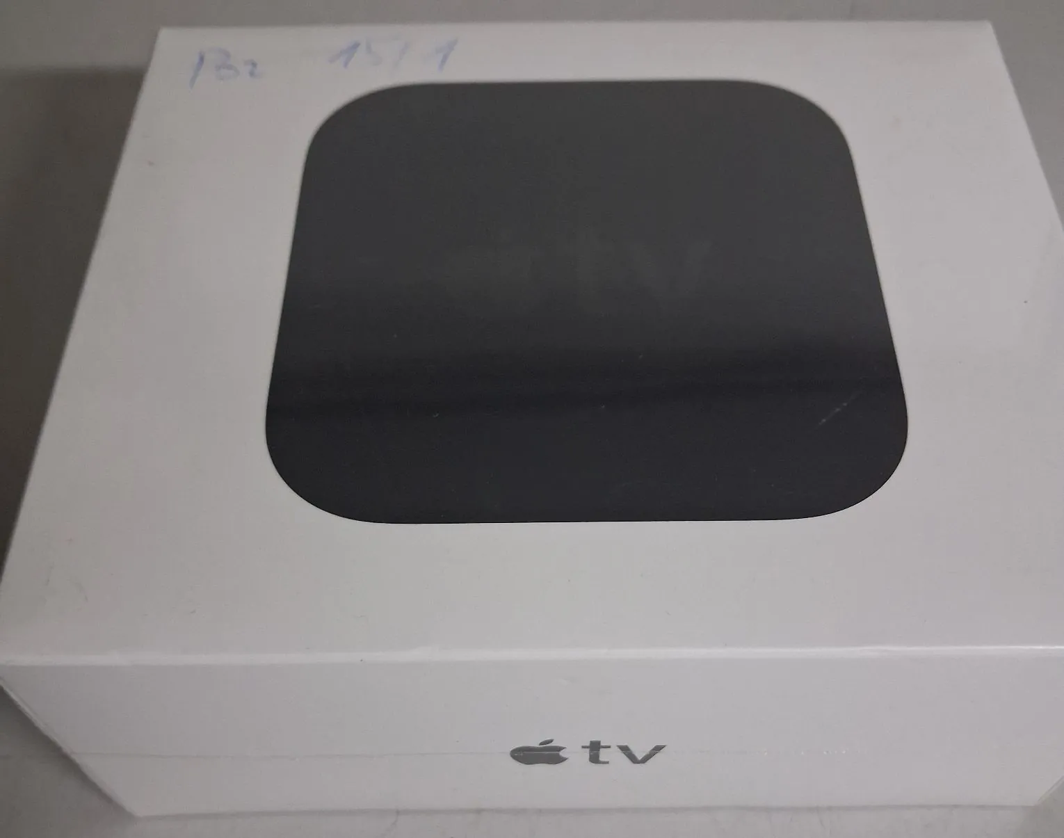 Białe pudełko z urządzeniem Apple TV. Na górze znajduje się duże, czarne, zaokrąglone okno, przez które widać produkt. Na dole pudełka widnieje logo Apple oraz napis „tv”. W lewym górnym rogu widoczne są odręczne zapiski długopisem: „B2 15/1”.