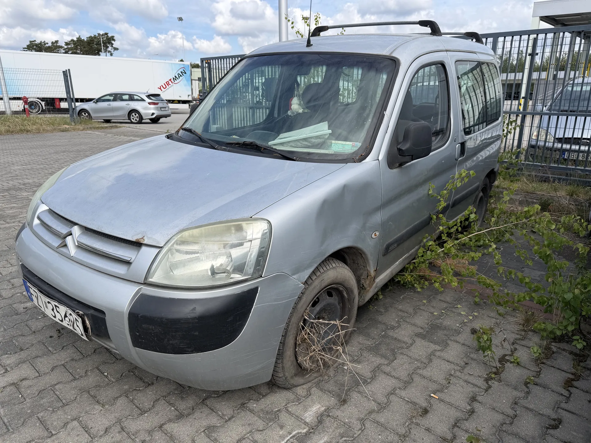 Srebrny samochód dostawczy marki Citroën z widocznym logo na przodzie, uszkodzonym przednim zderzakiem i oponą z suchą trawą wewnątrz. Tablica rejestracyjna z napisem EL 3569S. W tle biały naczepa z napisem YURTMAN, srebrny samochód osobowy, metalowe ogrodzenie, budynki i niebo z chmurami. Samochód stoi na brukowanej powierzchni z porośniętymi roślinami obszarami.