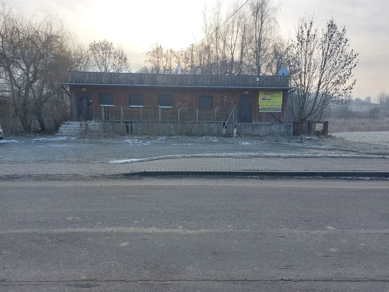 Zdjęcie przedstawiające parterowy, drewniany budynek o prostokątnym kształcie, znajdujący się przy drodze. Budynek ma ciemnobrązową elewację, kilka okien oraz wejście z małymi schodami po lewej stronie. Na ścianie budynku widoczne są graffiti oraz żółta tablica informacyjna z tekstem i numerami telefonów. Przed budynkiem znajduje się utwardzony plac, a na pierwszym planie asfaltowa droga. W tle widoczne są bezlistne drzewa pod jasnym, zimowym niebem.