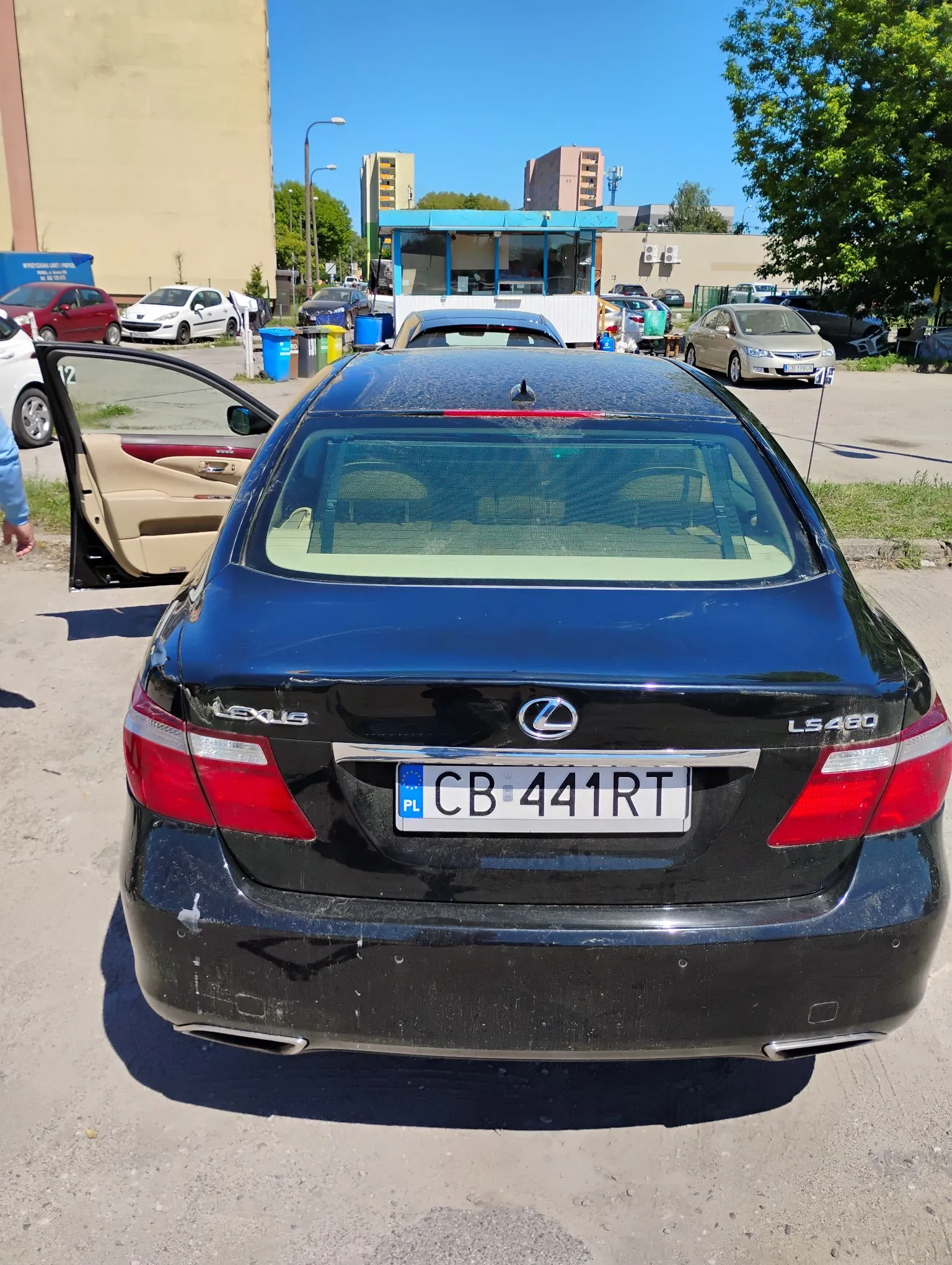 Czarny samochód marki Lexus LS 460 zaparkowany na placu. Widoczny jest tył pojazdu z polską tablicą rejestracyjną o numerze CB 441RT. W tle znajdują się inne zaparkowane samochody oraz budynki.