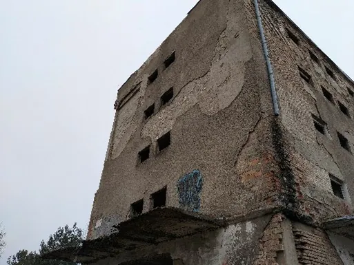 Zdjęcie przedstawiające narożnik starego, zniszczonego budynku z cegły i betonu. Elewacja jest mocno uszkodzona, z odpadającym tynkiem, odsłaniającym czerwoną cegłę. W ścianie widoczne są małe, prostokątne otwory okienne bez szyb. Na ścianie znajduje się fragment niebieskiego graffiti. Wzdłuż krawędzi budynku biegnie pionowa rura spustowa.