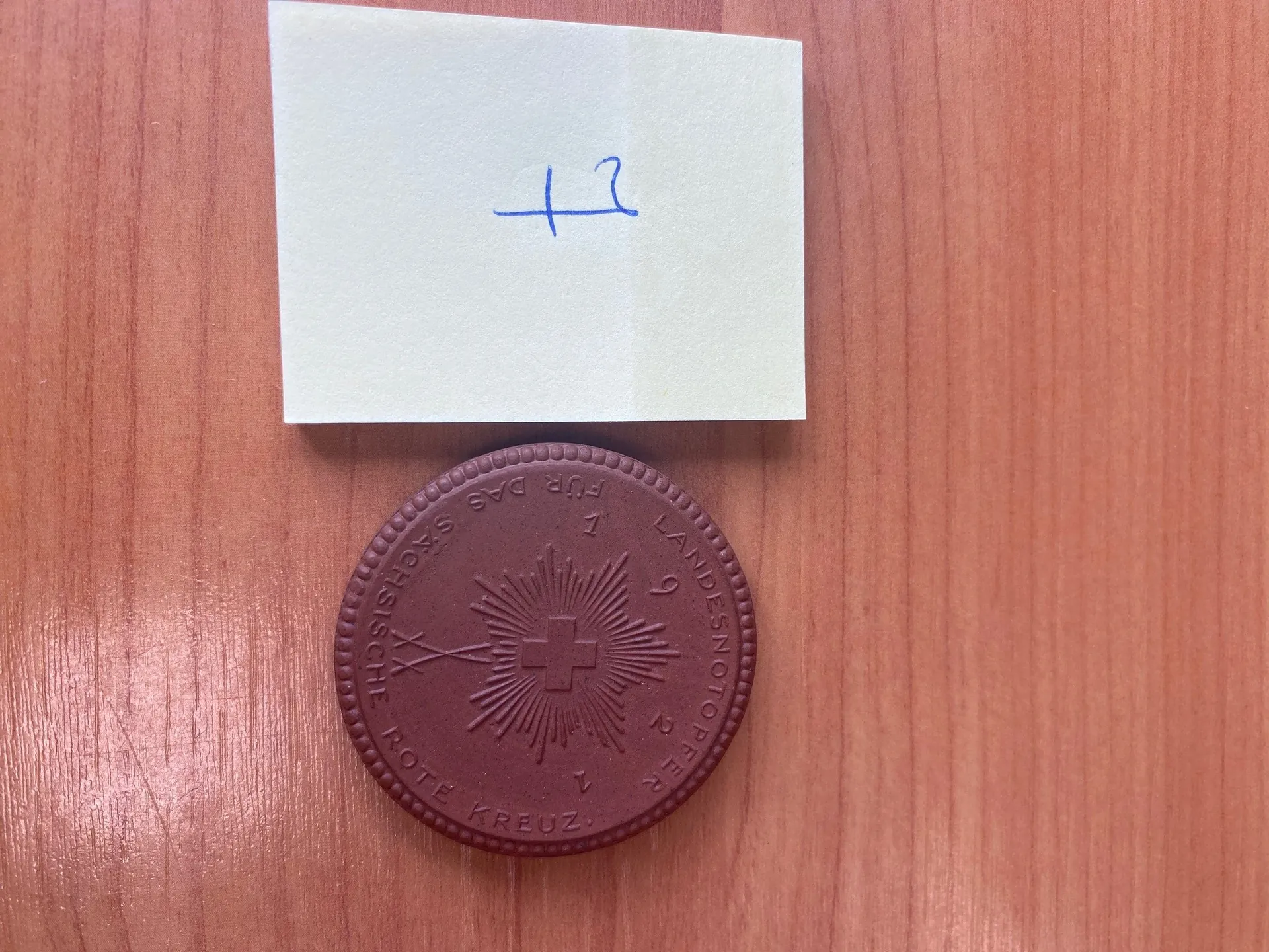 Na drewnianym tle leży biały kwadratowy kawałek papieru z niebieskim ręcznie napisanym '+2'. Poniżej papieru znajduje się brązowy okrągły medal z wypukłym krzyżem w centrum, promieniami rozchodzącymi się od krzyża oraz dwoma skrzyżowanymi mieczami po lewej stronie krzyża. Na medalu widoczny jest napis: 'FÜR DAS SÄCHSISCHE ROTE KREUZ', 'LANDESNOTOPFER 19'.
