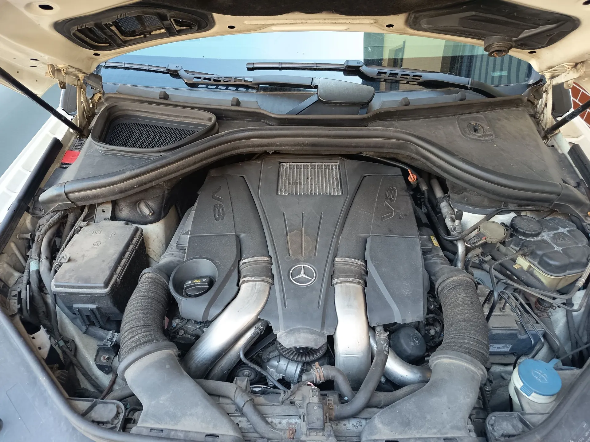 Widok z góry na komorę silnika samochodu z otwartą maską. W centrum znajduje się silnik V8 z plastikową osłoną, na której widnieje logo Mercedes-Benz oraz napisy 'V8' po obu stronach. Widoczne są srebrne rury dolotowe, czarne przewody, akumulator po lewej stronie oraz zbiornik płynu hamulcowego po prawej. Całość jest lekko zakurzona.
