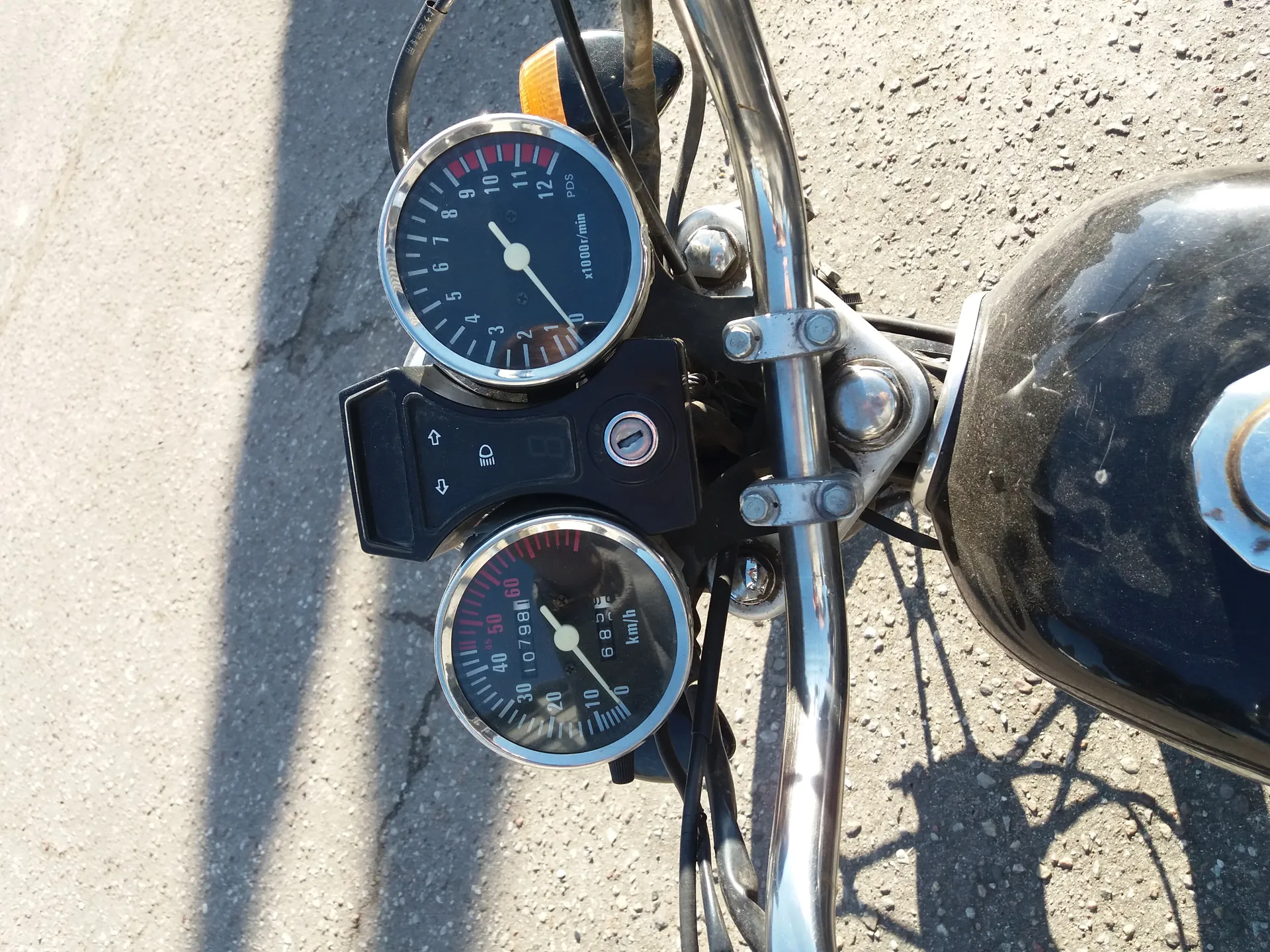 Zbliżenie na deskę rozdzielczą motocykla z dwoma okrągłymi wskaźnikami. Górnym jest tachometr z zakresem od 0 do 12 (oznaczony jako x1000r/min), a dolnym prędkościomierz w km/h do 60. Między nimi czarna panel z ikonami: strzałkami kierunkowymi, symbolem światła i otworem na klucz. Widoczne są również metalowe elementy kierownicy, przewody oraz czarna zbiornik paliwa. Tło stanowi asfaltowa droga z cieniami.