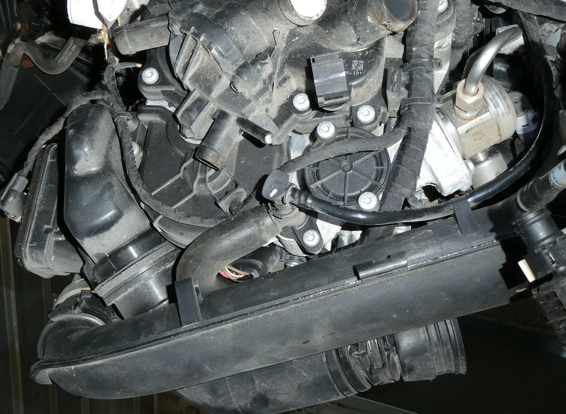 Zbliżenie na zakurzone elementy silnika samochodu, widoczne są czarne plastikowe obudowy, przewody, wąż dolotowy oraz metalowe śruby i złącza.
