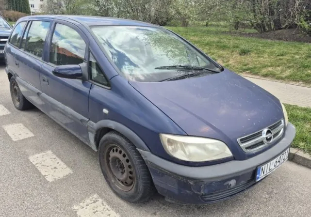 Niebieski samochód osobowy marki Opel, minivan, zaparkowany na ulicy. Widoczna tablica rejestracyjna NTL 547Z. Samochód ma brudną karoserię, przednie światła i logo Opel na masce. W tle trawnik, krzewy i chodnik. Na jezdni białe znakowanie.