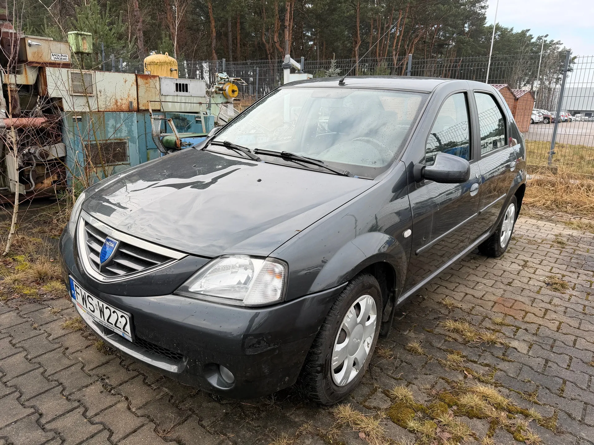 Ciemnoszary samochód osobowy marki Dacia zaparkowany na brukowanej powierzchni. Widoczna tablica rejestracyjna FWS W222. W tle stoją zardzewiałe maszyny przemysłowe, metalowe ogrodzenie, drzewa oraz budynki. Na ziemi rośnie sucha trawa i mchy. Samochód ma srebrne felgi i niebieski emblemat marki na masce.