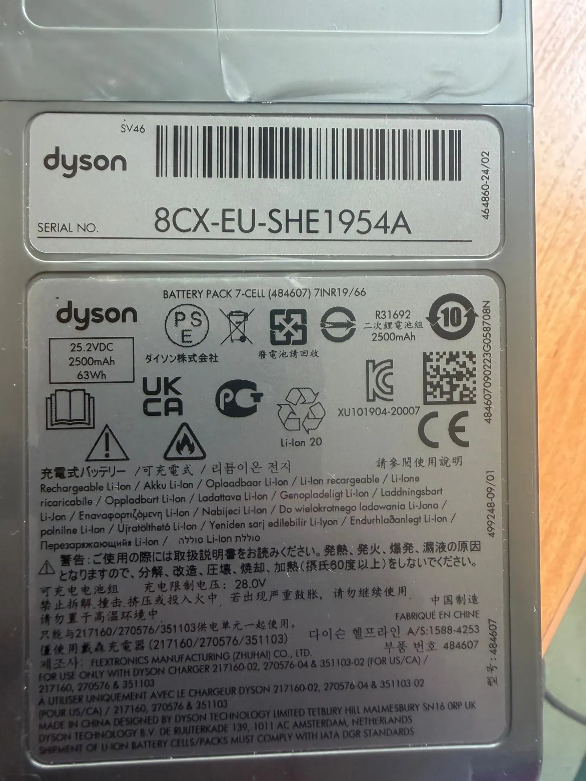 Zdjęcie etykiety technicznej baterii marki Dyson. Na górze znajduje się logo 'dyson' oraz numer seryjny '8CX-EU-SHE1954A'. Poniżej widnieje napis 'BATTERY PACK 7-CELL (484607) 7INR19/66'. Etykieta zawiera specyfikację techniczną: 25.2VDC, 2500mAh, 63Wh. Widoczne są liczne symbole certyfikacji, w tym CE, UKCA, znak recyklingu oraz ostrzeżenia w wielu językach. W dolnej części znajduje się tekst informujący o producentie: 'MADE IN CHINA DESIGNED BY DYSON TECHNOLOGY LIMITED'.
