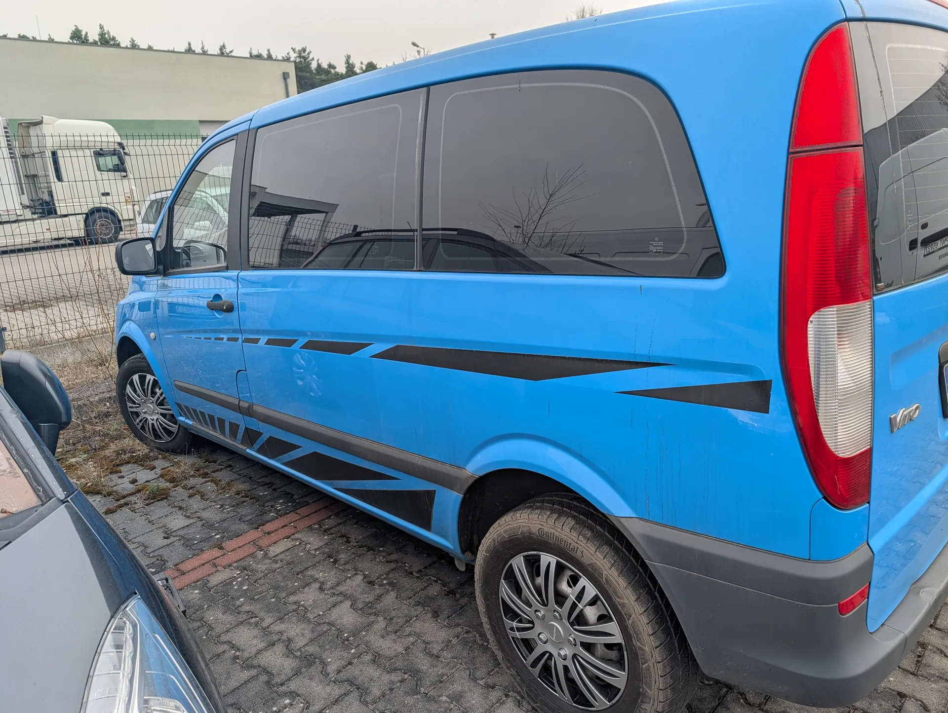Niebieski samochód dostawczy Mercedes-Benz Vito z czarnymi dekoracyjnymi pasami na boku, zaparkowany na brukowanej powierzchni. Widoczne są czerwone światła tylne, srebrne felgi oraz napis "Vito" na tylnej części pojazdu. W tle metalowe ogrodzenie, biały ciężarówka, budynek i drzewa. Częściowo widoczny szary samochód na pierwszym planie po lewej stronie.