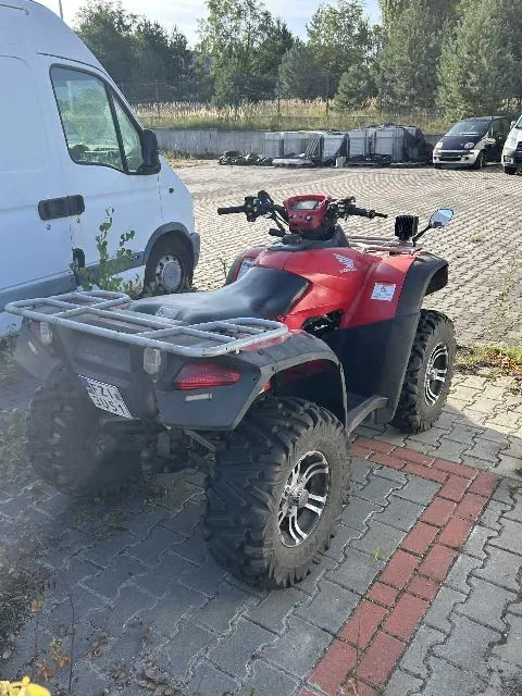 Czerwony quad marki Honda zaparkowany na kostce brukowej obok białego vana. Pojazd posiada srebrne bagażniki z przodu i z tyłu oraz duże, terenowe opony. Z tyłu widoczna jest tablica rejestracyjna o numerze PZI 3D51.