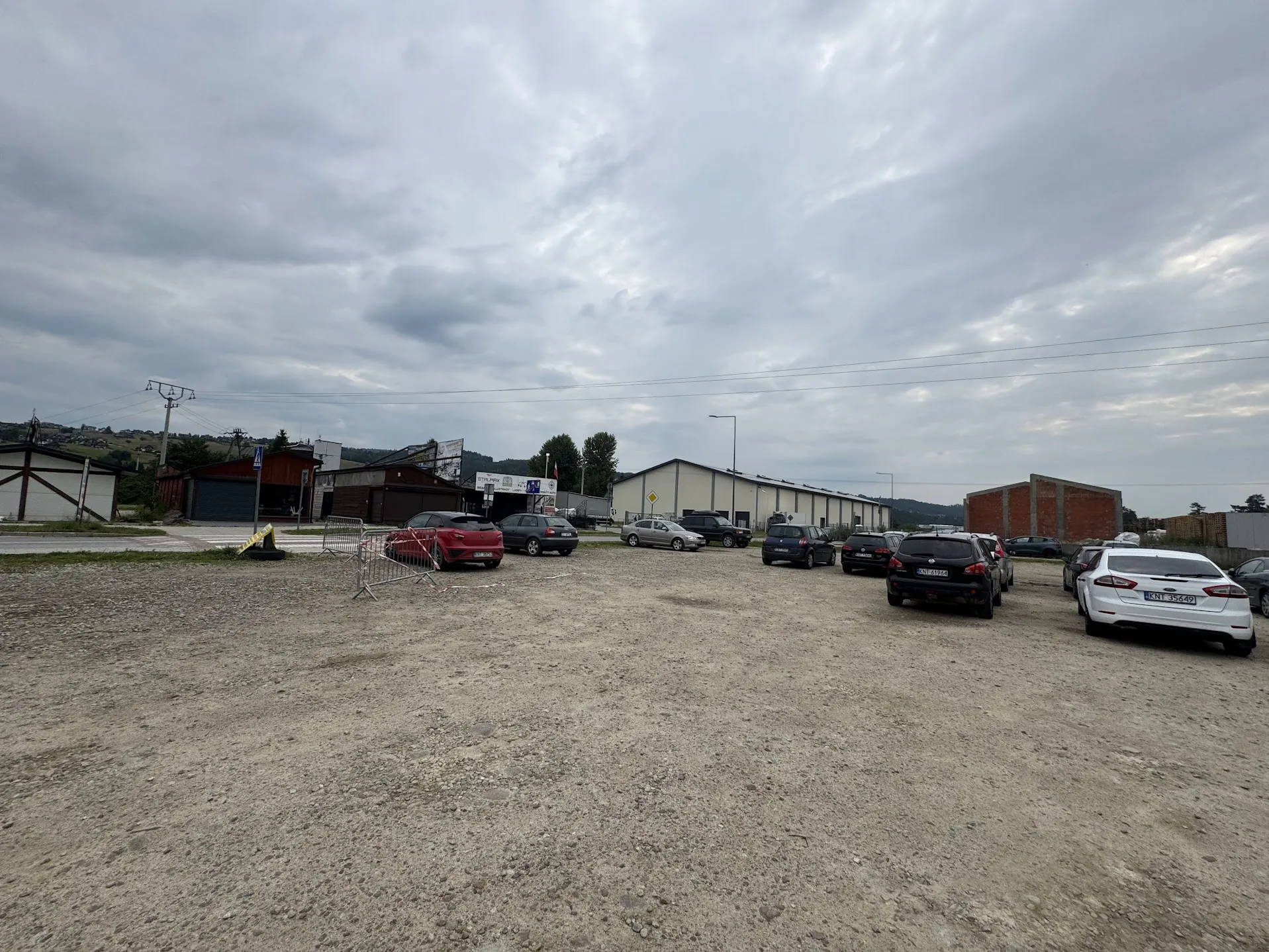 Gruntowy parking z kilkoma zaparkowanymi samochodami, w tym czerwonym, czarnym i białym. W tle budynki przemysłowe, w tym biały hangar i czerwona ceglaną struktura. Widoczne są znaki drogowe, barierki i słup z liniami energetycznymi. Niebo jest zachmurzone. Na białym samochodzie tablica rejestracyjna KNT 35449. W oddali wzgórza i drzewa.