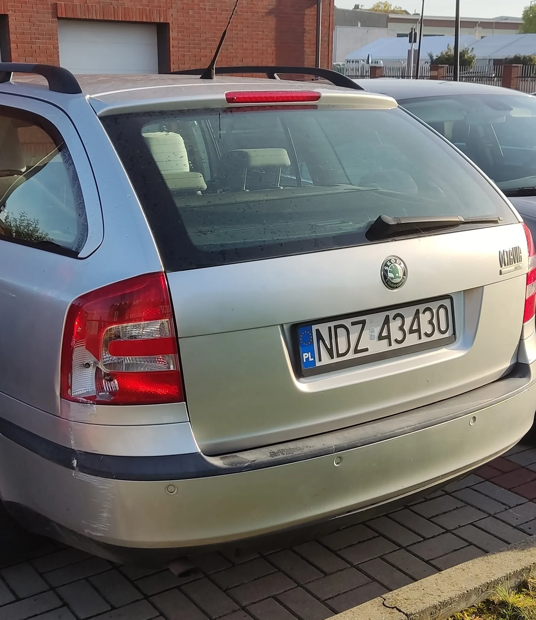 Tylny widok srebrnego samochodu marki Skoda Octavia Combi zaparkowanego na kostce brukowej. Samochód ma polską tablicę rejestracyjną o numerze NDZ 43430. Na tylnym zderzaku po lewej stronie widoczne są zarysowania.