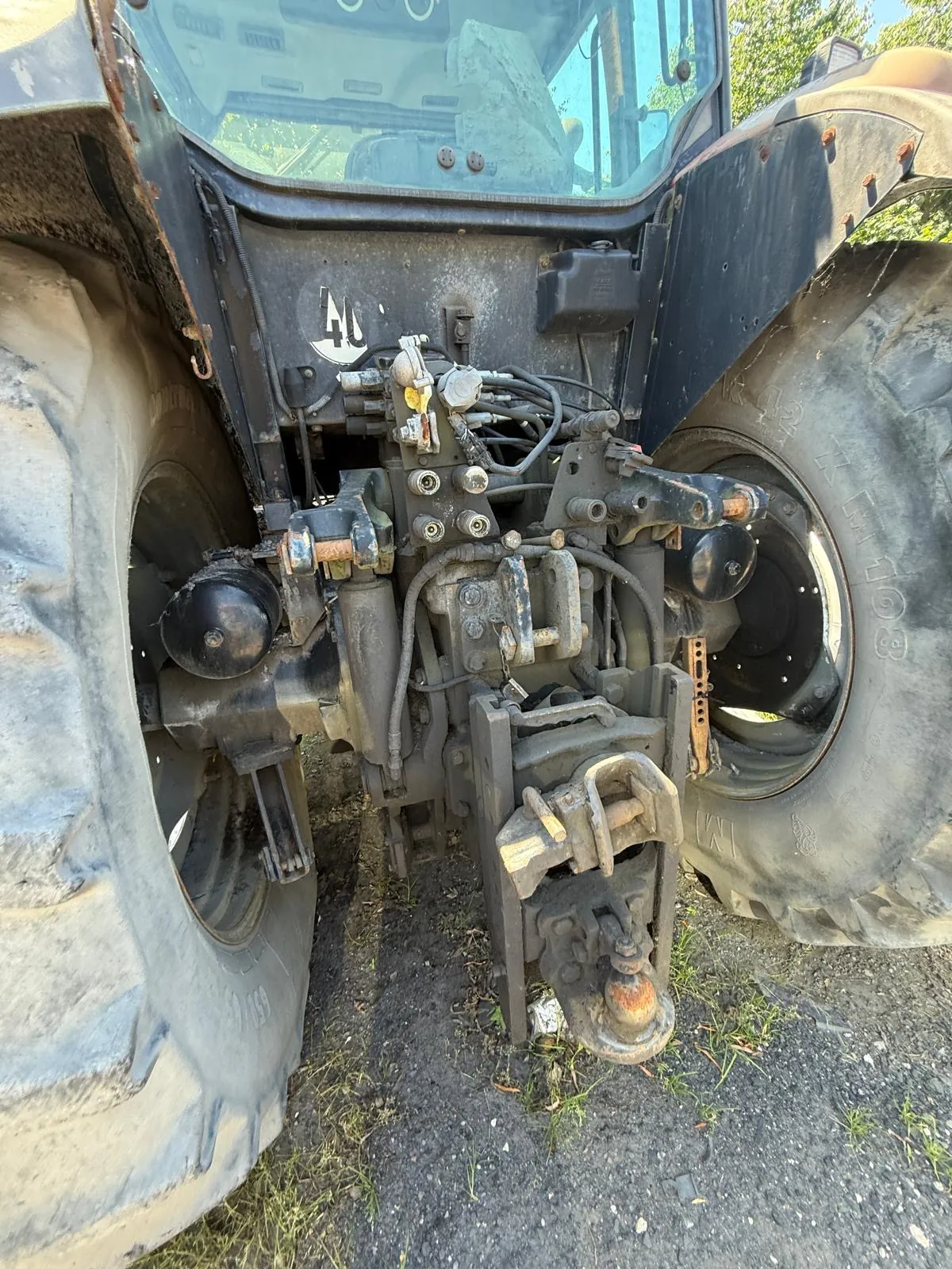 Zbliżenie na tylną część traktora, ukazujące układ hydrauliczny, sprzęg sprzęgowy oraz duże, brudne opony z obu stron. Widoczne są metalowe elementy konstrukcyjne, przewody hydrauliczne i śruby, na niektórych częściach widoczna jest rdza.