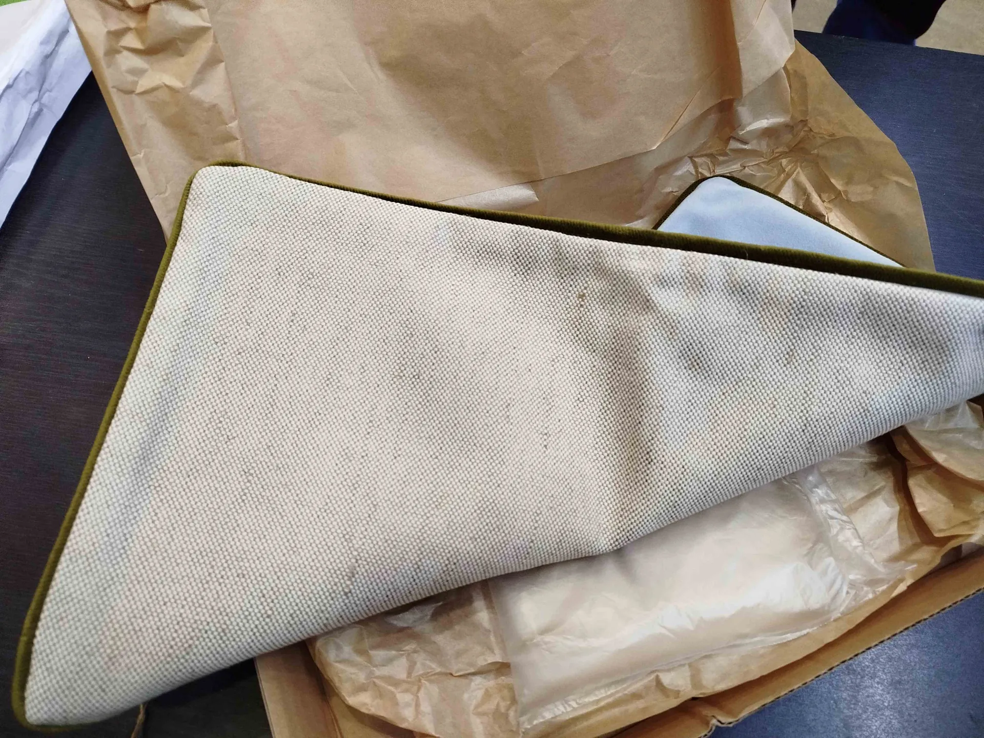 Zdjęcie przedstawia składany przedmiot z tkaniny w kolorze beżowym z zielonym wykończeniem brzegów. Przedmiot leży na brązowym papierze pakowym wewnątrz kartonowego pudełka. Pod tkaniną widoczne są przezroczyste opakowanie z plastiku oraz dodatkowe warstwy papieru pakowego. Tło stanowi ciemna powierzchnia stołu i fragment białego materiału po lewej stronie.