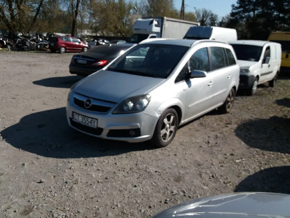 Srebrny samochód Opel z tablicą rejestracyjną ACT 3554Y na pierwszym planie, zaparkowany na żwirowym placu. W tle widoczne są inne pojazdy: czarny samochód, biały furgon, czerwony samochód osobowy oraz ciężarówki. Tło stanowią drzewa i jasne niebo. Podłoga jest nierówna, z widocznymi kamieniami i cieniami od pojazdów.