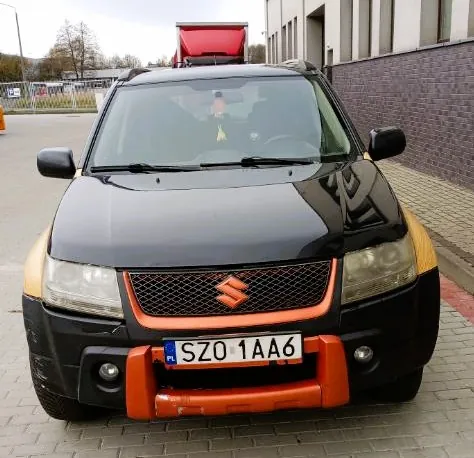Czarny samochód marki Suzuki widziany od przodu. Pojazd posiada pomarańczowe elementy wykończenia na przednim zderzaku oraz nadkolach. Na masce widoczne jest logo Suzuki. Tablica rejestracyjna ma numer SZO 1AA6. Samochód zaparkowany jest na kostce brukowej obok budynku z ciemnobrązową ceglaną ścianą.