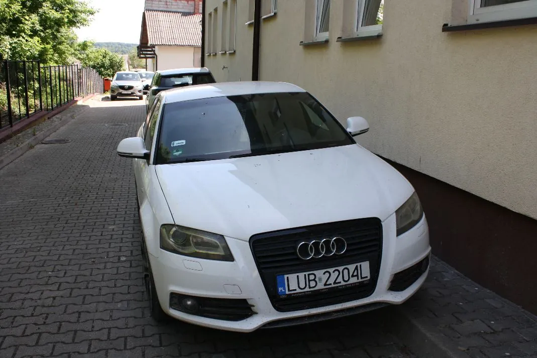 Biały samochód marki Audi zaparkowany na brukowanej alejce obok jasnej ściany budynku. W tle widoczny jest kolejny biały samochód oraz fragmenty zieleni i dachu budynku. Samochód na pierwszym planie ma tablicę rejestracyjną o treści LUB 2204L.
