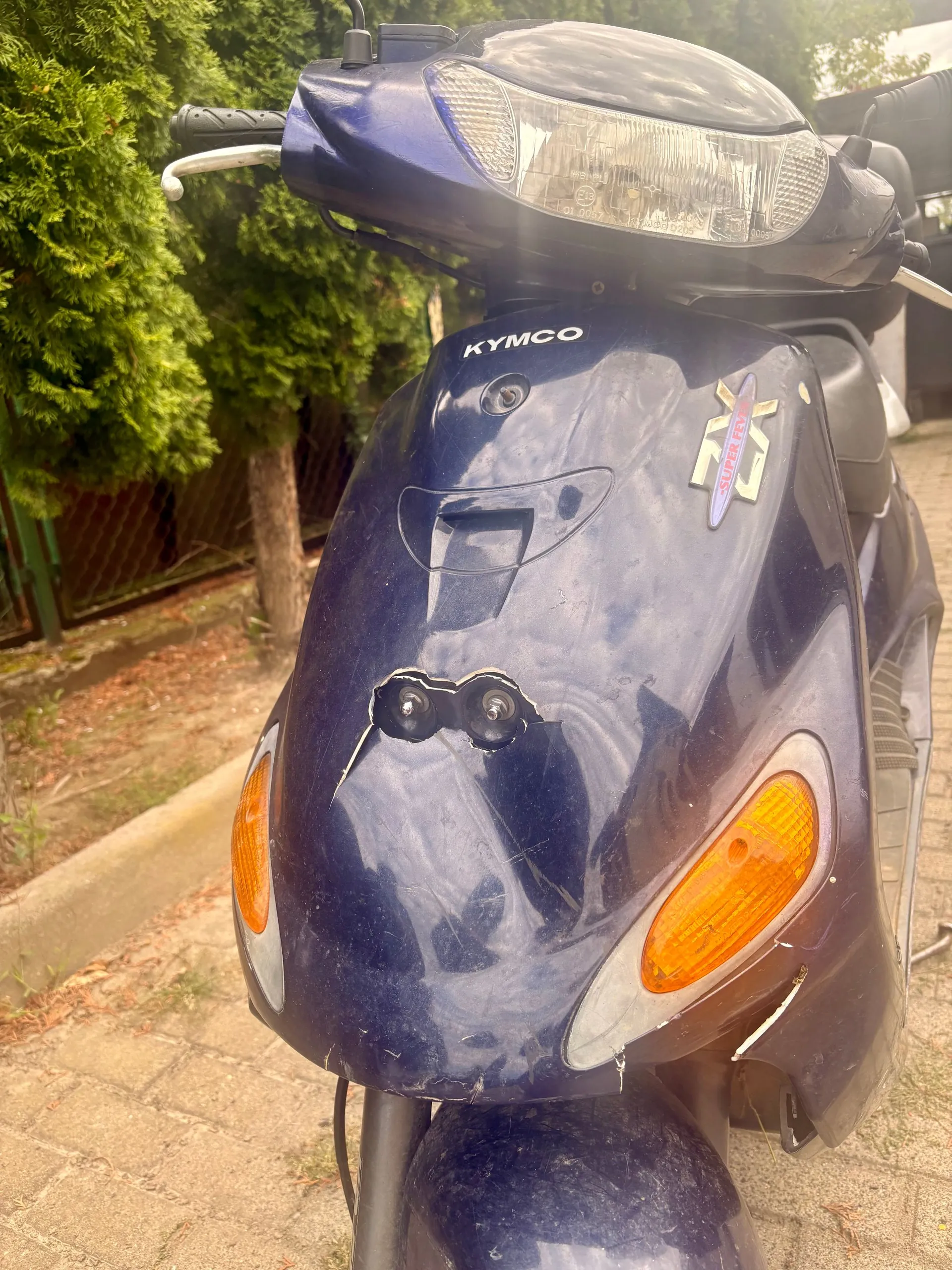 Zbliżenie na przód granatowego skutera marki Kymco. Widoczna jest przednia lampa, kierownica oraz owiewka z napisem KYMCO i naklejką z napisem SUPER FUN. Na przedniej obudowie widoczne są zarysowania oraz dwa otwory montażowe po zdjętej tablicy rejestracyjnej. Po bokach znajdują się pomarańczowe kierunkowskazy. W tle widać zielone krzewy i fragment chodnika.