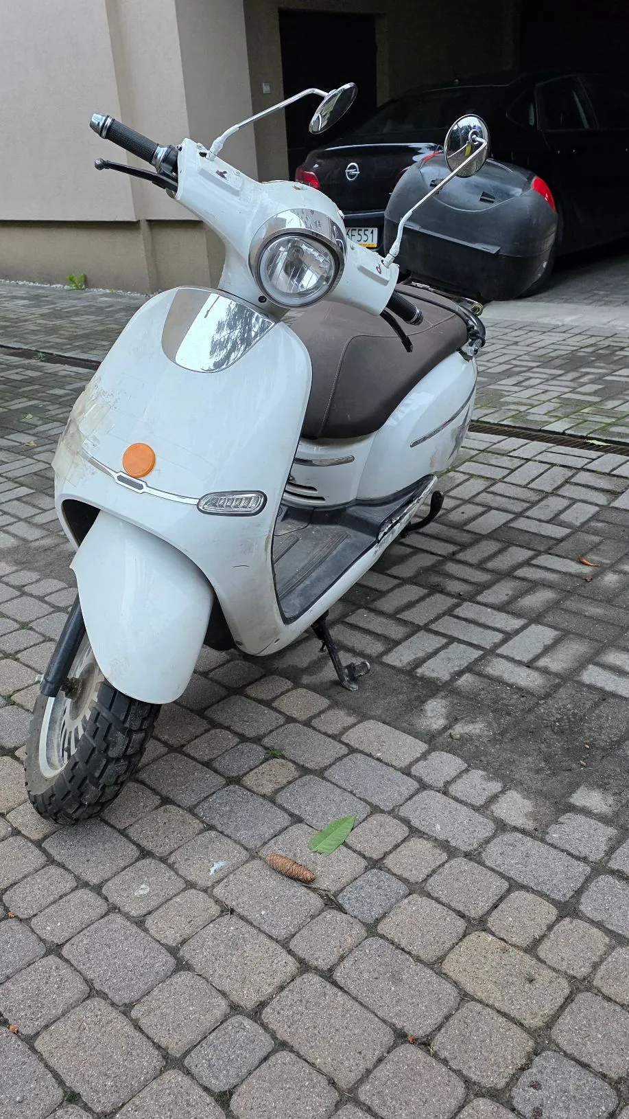 Biały skuter zaparkowany na kostce brukowej. Pojazd ma brązowe siedzenie i jest ustawiony pod kątem do widza. W tle widoczny jest czarny samochód osobowy zaparkowany przed budynkiem.