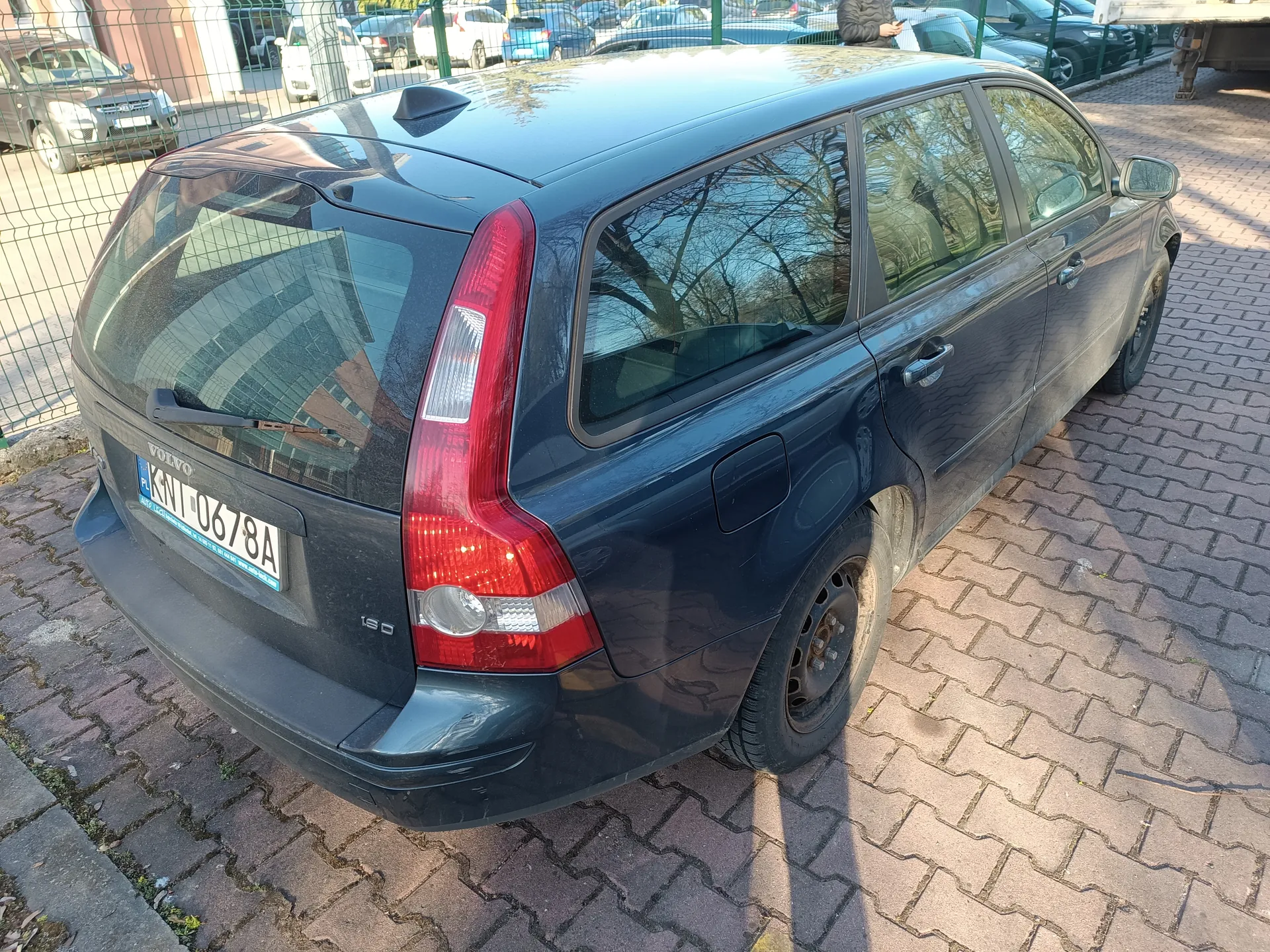Ciemnoniebieski samochód osobowy typu kombi marki Volvo z tablicą rejestracyjną KWI 0678A, zaparkowany na brukowanej powierzchni. Widok z tyłu i boku. Na tylnej szybie widoczny napis 'VOLVO' oraz oznaczenie '120' na lewej stronie bagażnika. W tle metalowe ogrodzenie, inne zaparkowane samochody oraz budynki.