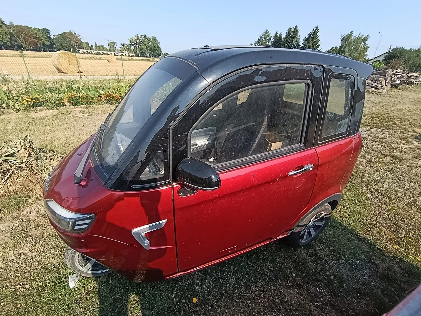 Czerwony, kompaktowy samochód elektryczny o zaokrąglonym kształcie, zaparkowany na trawiastym terenie. Pojazd ma czarny dach i czarne ramki okien. W tle widoczne jest pole z belami siana oraz drzewa pod jasnym, niebieskim niebem.