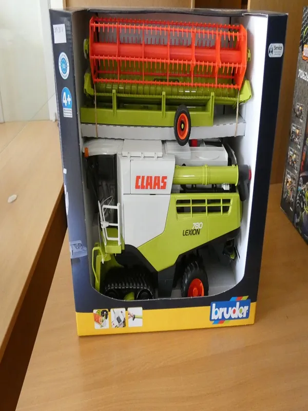 Zabawkowy model kombajnu CLAAS 780 LEXION w opakowaniu marki bruder. Opakowanie czarno-żółte z przezroczystym okienkiem, na którym widoczny zielono-biały model z pomarańczowymi elementami. Na opakowaniu widnieje napis '4+' oraz logo 'bruder' w dolnej części. Model umieszczony na drewnianym stole, w tle fragment innego opakowania.