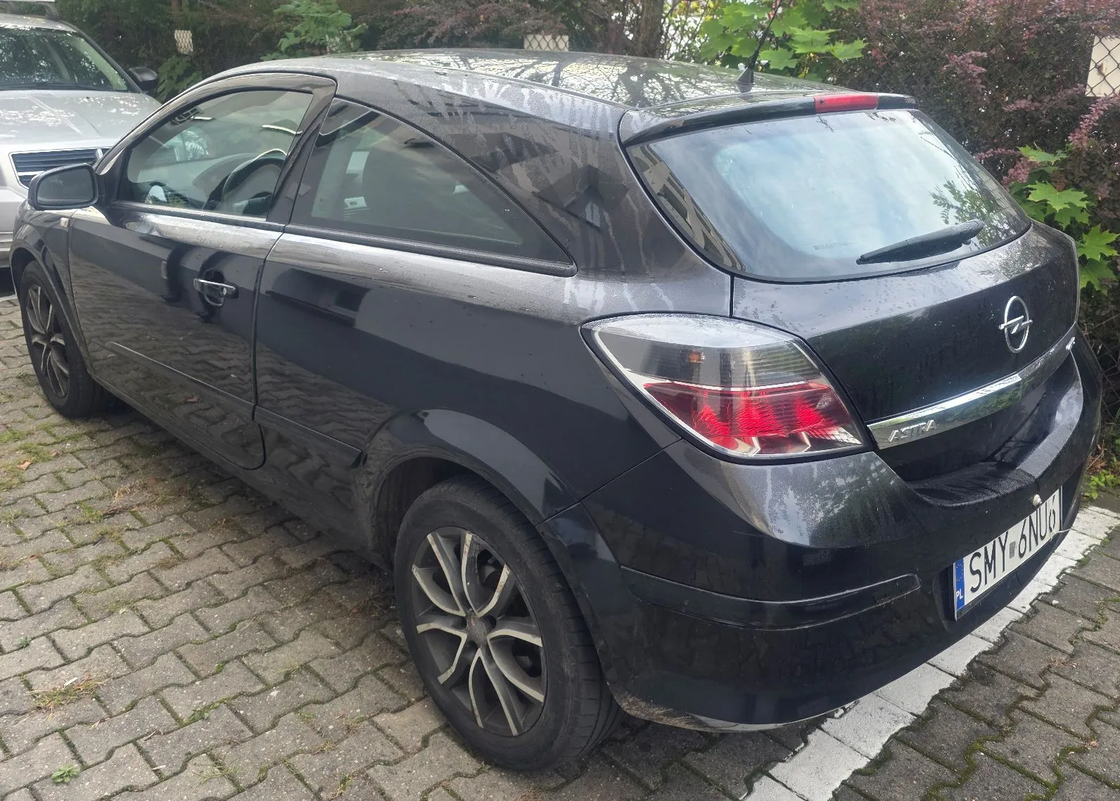 Czarny samochód marki Opel Astra zaparkowany na kostce brukowej. Widok z tyłu i z boku pojazdu. Samochód ma tablicę rejestracyjną o numerze SMY 6NU6.