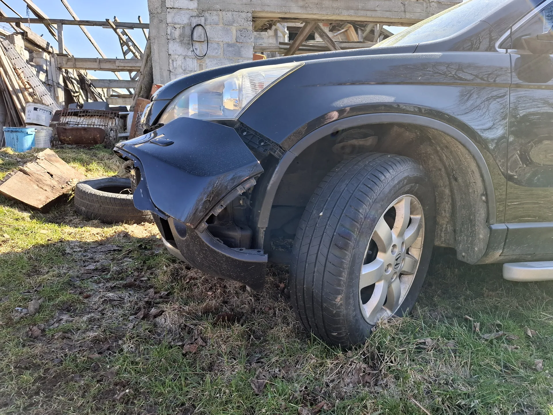 Czarny SUV z uszkodzonym przodem, z pękniętym i zwisającym zderzakiem, zaparkowany na trawie z suchymi liśćmi. W tle betonowa konstrukcja budowlana z drewnianymi belkami, sterty materiałów, niebieskim i białym wiadrem oraz starym oponą. Jasne niebo.