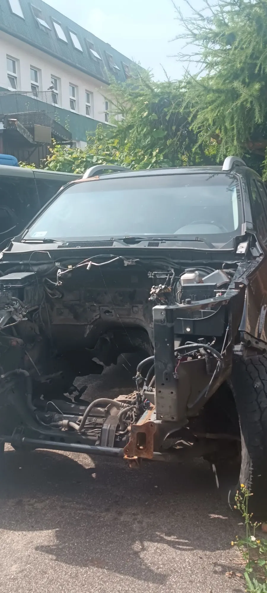 Zdjęcie przedstawiające przód czarnego samochodu typu SUV, który jest w stanie rozbiórki lub po poważnej awarii. Brakuje maski, przedniego zderzaka, reflektorów oraz grilla, co odsłania wewnętrzne elementy konstrukcyjne, wiązki elektryczne i podwozie. Pojazd stoi na asfaltowej nawierzchni na tle zielonych krzewów i białego budynku z wielospadowym dachem.