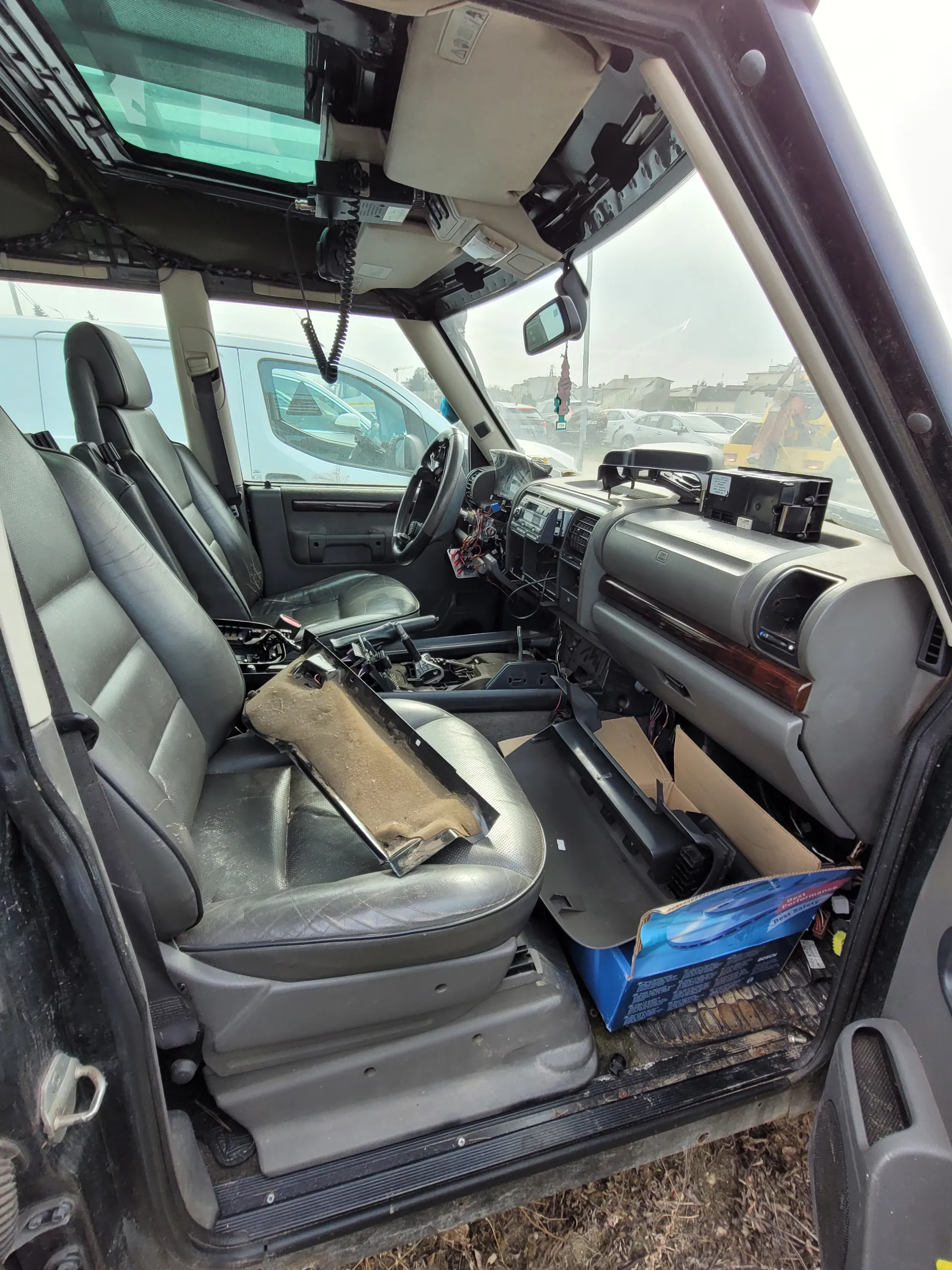 Wnętrze samochodu z otwartymi drzwiami kierowcy. Szare skórzane fotele, na siedzeniu pasażera niebieskie pudełko i karton. Deska rozdzielcza częściowo rozebrana z widocznymi przewodami. Przez przednią szybę widoczny inny pojazd i budynki. Poza samochodem sucha trawa i ziemia.