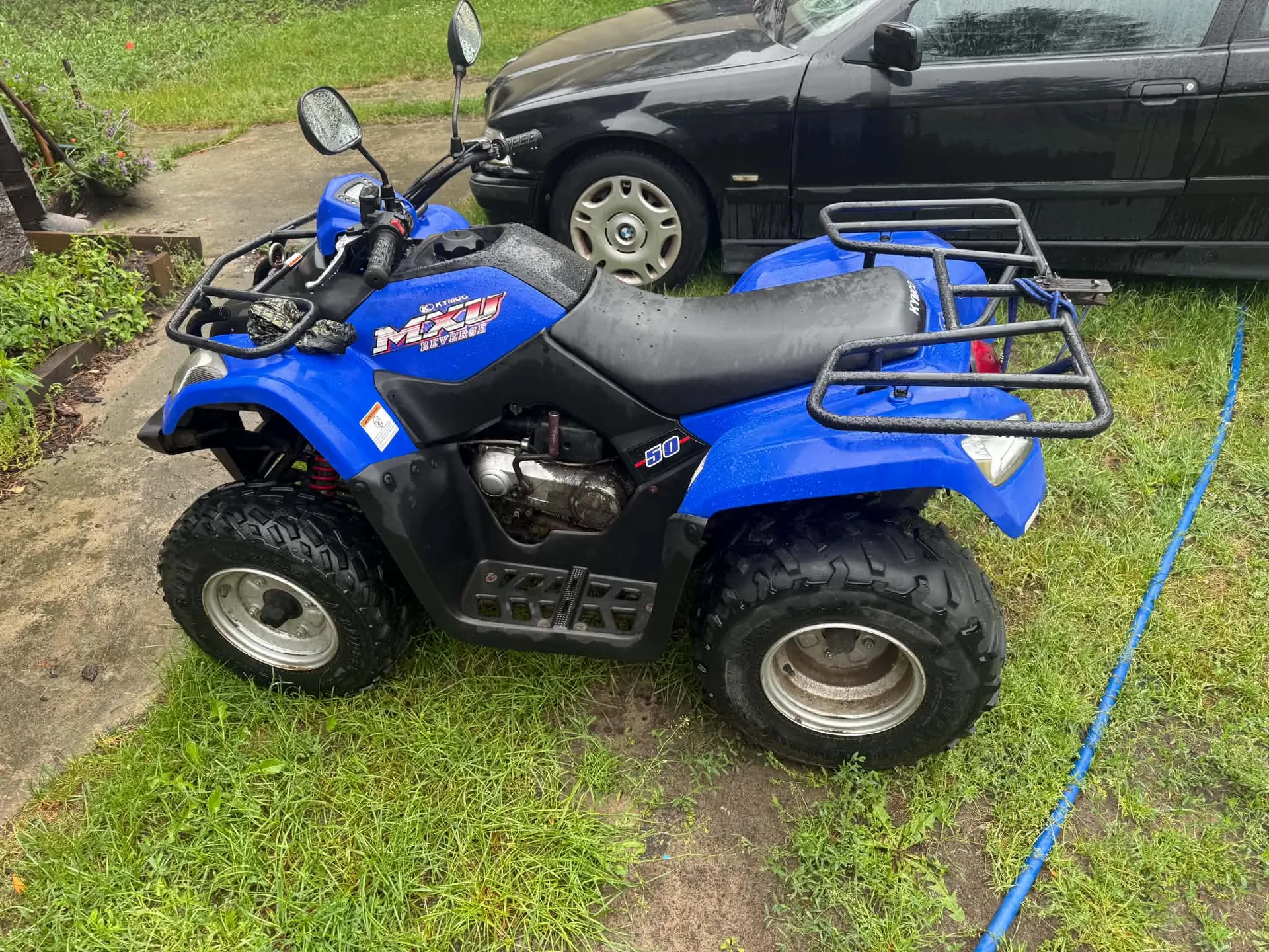 Niebieski quad marki MXU stojący na trawie obok czarnego samochodu. Pojazd posiada czarne opony z grubym bieżnikiem, srebrne felgi, przedni i tylny bagażnik oraz lusterka wsteczne. Na boku widnieje napis 'MXU' oraz liczba '50'.