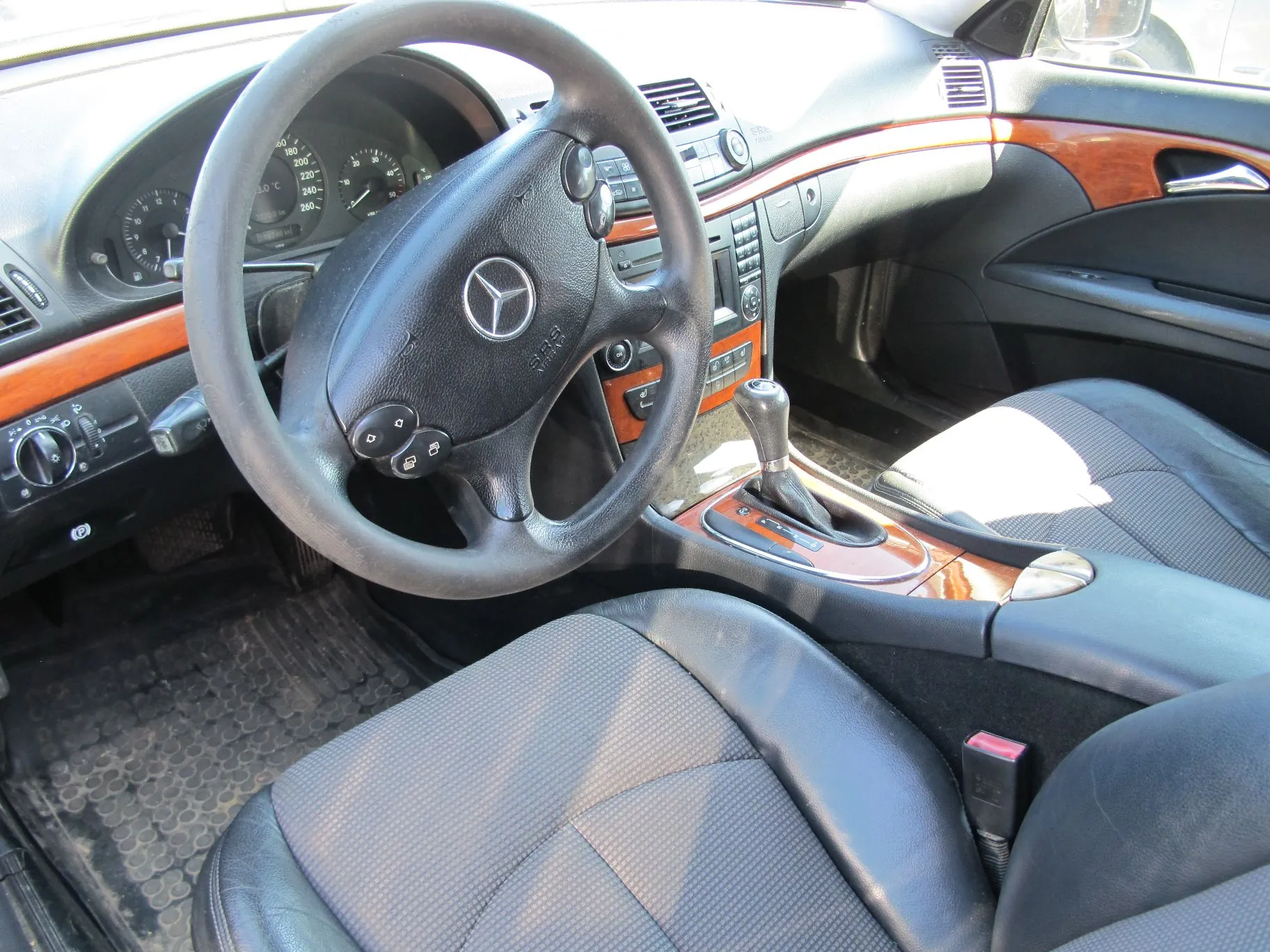 Wnętrze samochodu marki Mercedes-Benz z perspektywy kierowcy. Widoczna jest czarna skórzana kierownica z logo marki na środku, deska rozdzielcza z drewnianymi wstawkami, zegary wskaźników oraz czarne skórzane fotele. W centralnej konsoli znajduje się dźwignia zmiany biegów.