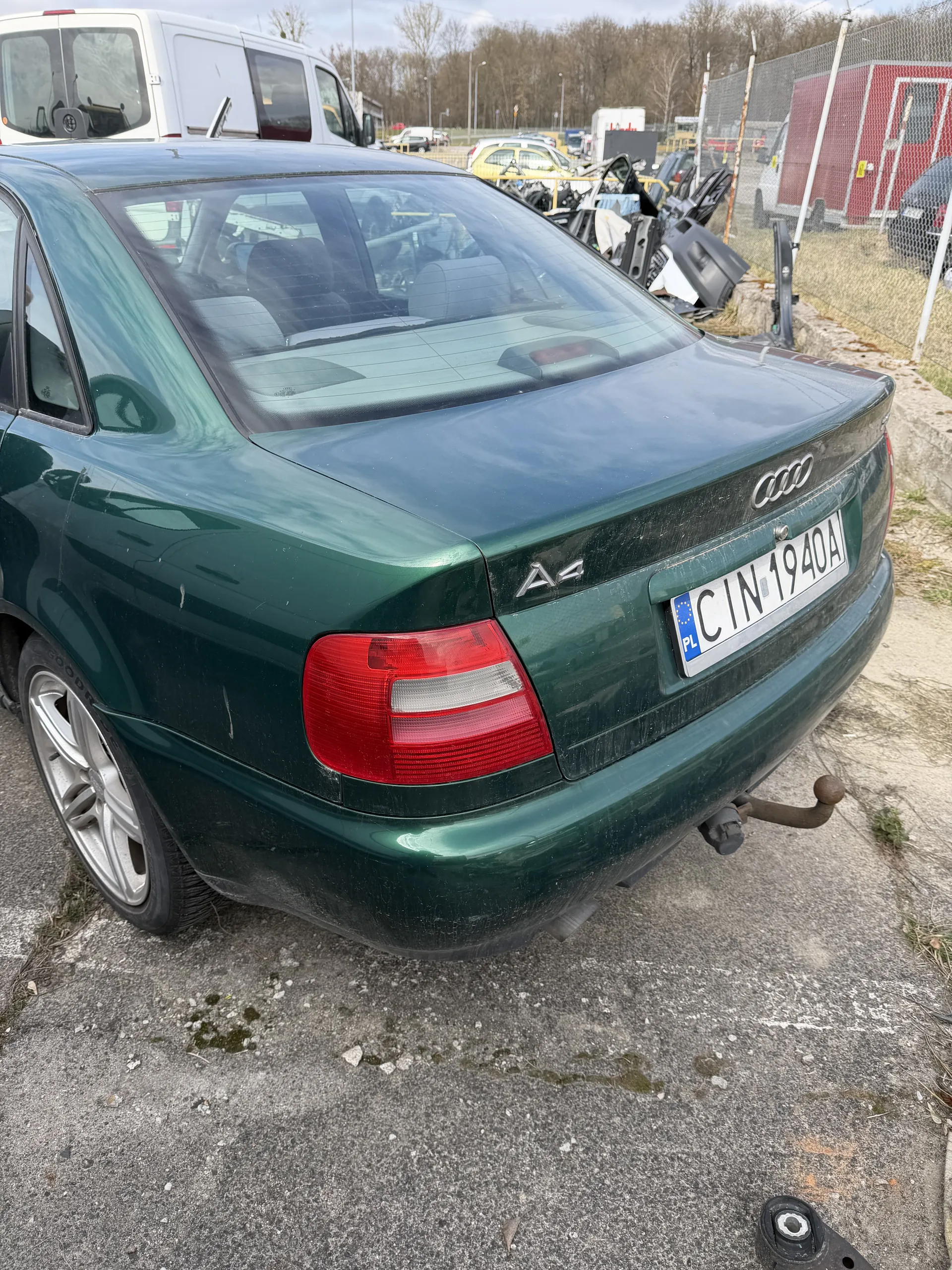 Z tyłu zielony samochód marki Audi A4 z tablicą rejestracyjną CIN 1904 PL. Samochód stoi na asfalcie, w tle widoczne są inne pojazdy, części samochodowe oraz czerwona budowla. Na asfalcie znajdują się plamy i kamienie.