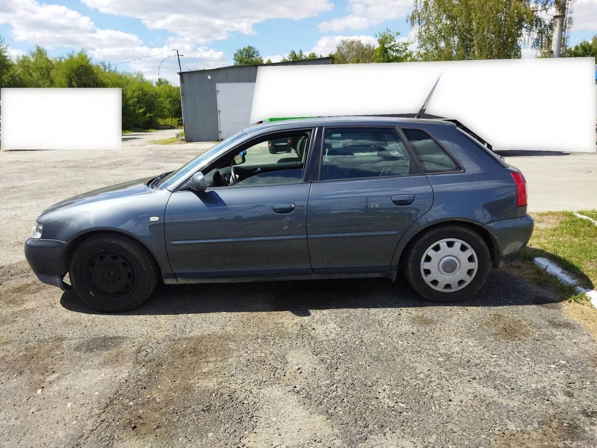 Szary samochód osobowy typu hatchback zaparkowany bokiem na żwirowym placu. W tle widoczny jest szary budynek gospodarczy, zielone drzewa oraz niebo z białymi chmurami.