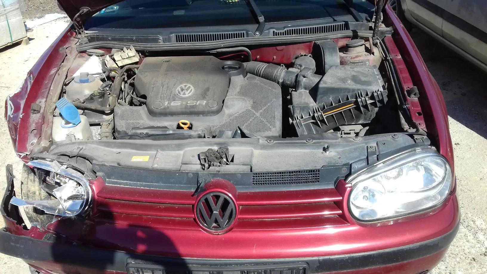 Widok z góry na otwartą maskę czerwonego samochodu marki Volkswagen. Widoczny jest silnik z plastikową osłoną z logo VW i napisem '1.6 SR'. Przednia część samochodu jest uszkodzona, szczególnie po lewej stronie, gdzie reflektor jest rozbity. Na środku grilla znajduje się duże logo Volkswagena.