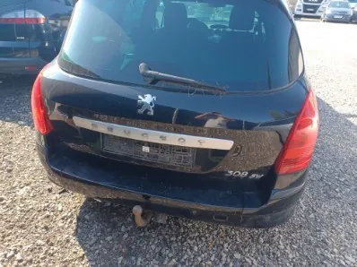 Tylny widok czarnego samochodu marki Peugeot model 308 SW. Na klapie bagażnika widoczne jest logo lwa oraz napis PEUGEOT na srebrnej listwie. Po prawej stronie znajduje się oznaczenie modelu 308 SW. Samochód posiada hak holowniczy i jest zaparkowany na żwirowym podłożu.
