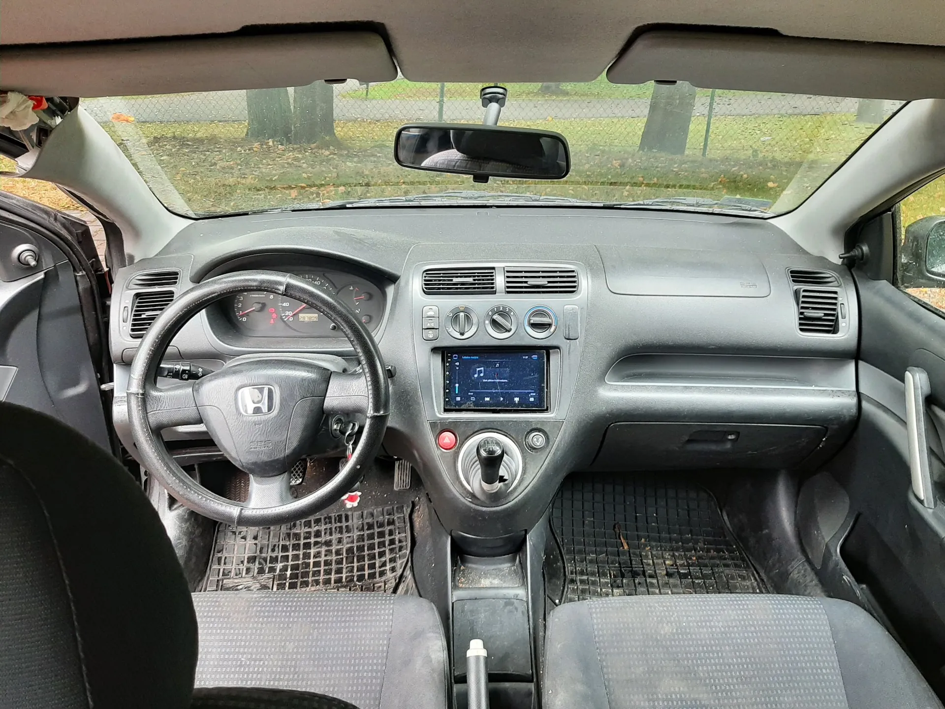 Wnętrze samochodu marki Honda widziane z perspektywy tylnego siedzenia. Widoczna jest deska rozdzielcza w kolorze ciemnoszarym, kierownica z logo Hondy, licznik, centralna konsola z zamontowanym ekranem multimedialnym oraz dwa przednie fotele z ciemną tapicerką. Na podłodze znajdują się gumowe maty. Przez przednią szybę widoczny jest fragment zielonego terenu z drzewami.