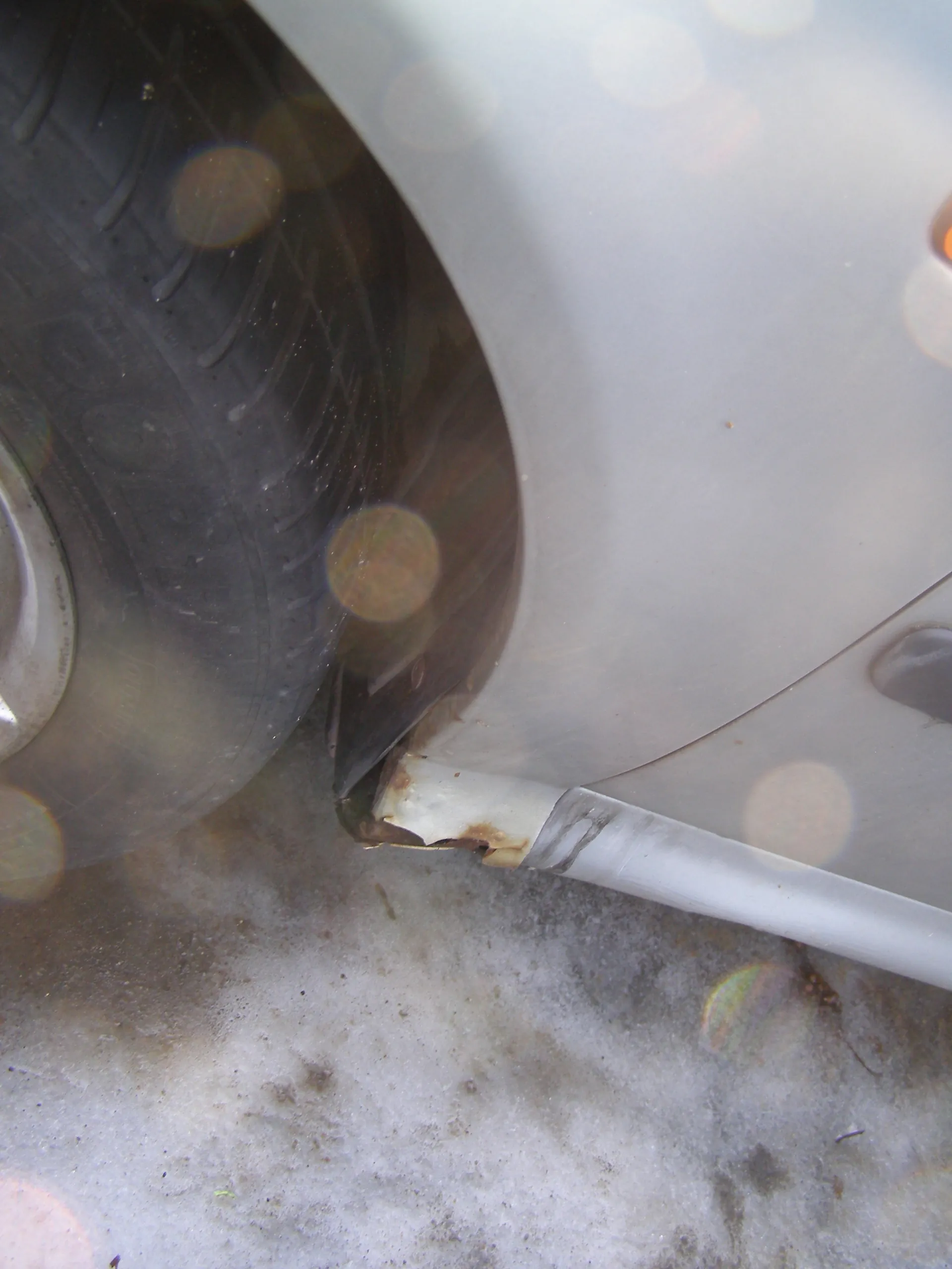 Zbliżenie na przednią część koła srebrnego samochodu. Widoczna opona z wyraźnymi śladami, rdza na białym plastikowym wykończeniu nadkola oraz śnieg z brudnymi plamami na powierzchni ziemi. Na obrazie występują efekty świetlne w postaci okrągłych plam od refleksów słońca.