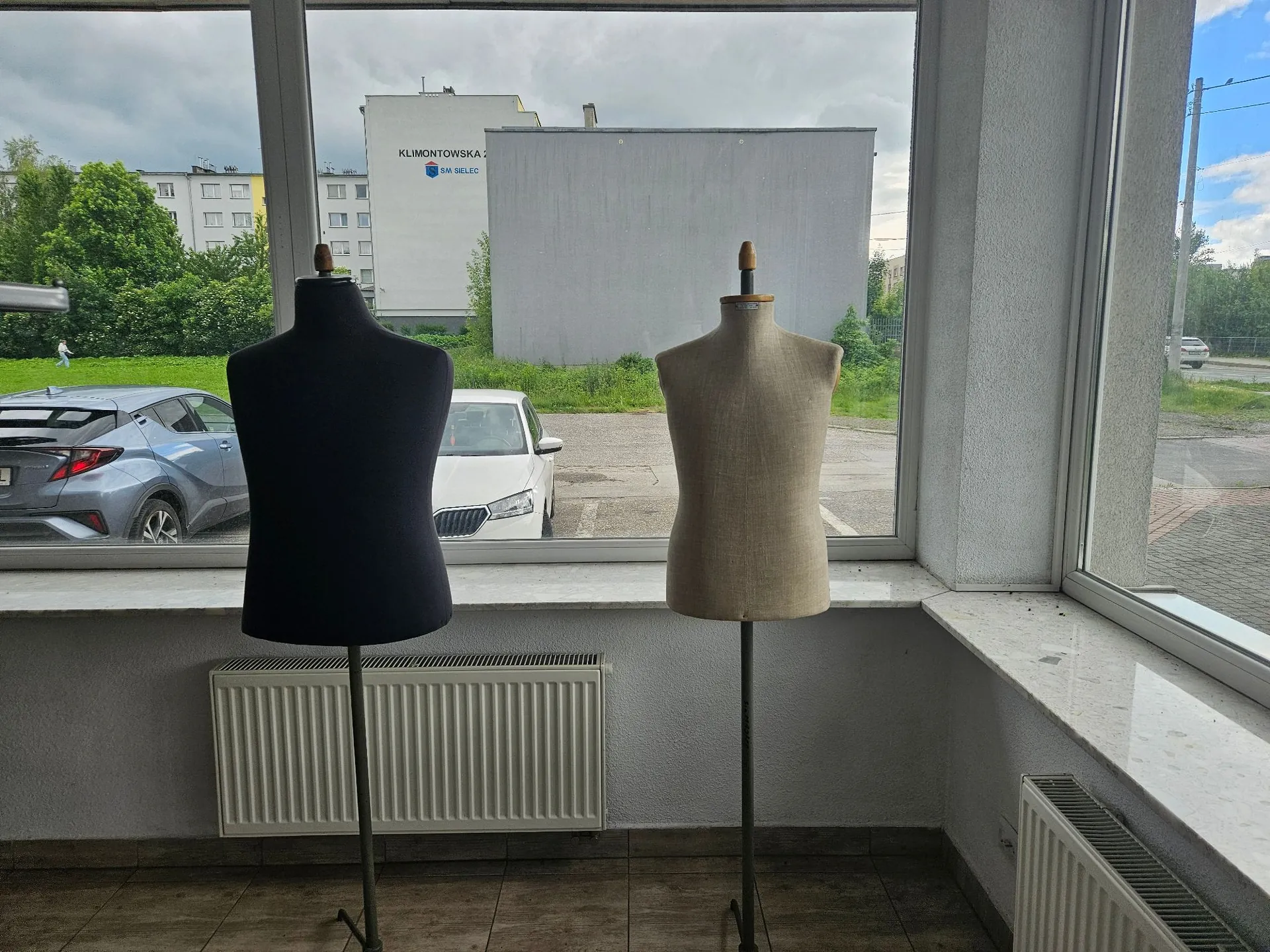 Wnętrze pomieszczenia z widokiem przez duże okno na zewnątrz. Wewnątrz stoją dwa manekiny krawieckie na statywach: jeden w kolorze ciemnogranatowym po lewej i jeden w kolorze beżowym po prawej. Pod oknem znajduje się biały grzejnik panelowy. Za oknem widoczny jest parking z zaparkowanym niebieskim i białym samochodem, zielona roślinność oraz jasny budynek z napisem 'KLIMONTOWSKA 2 SM SELEC'.