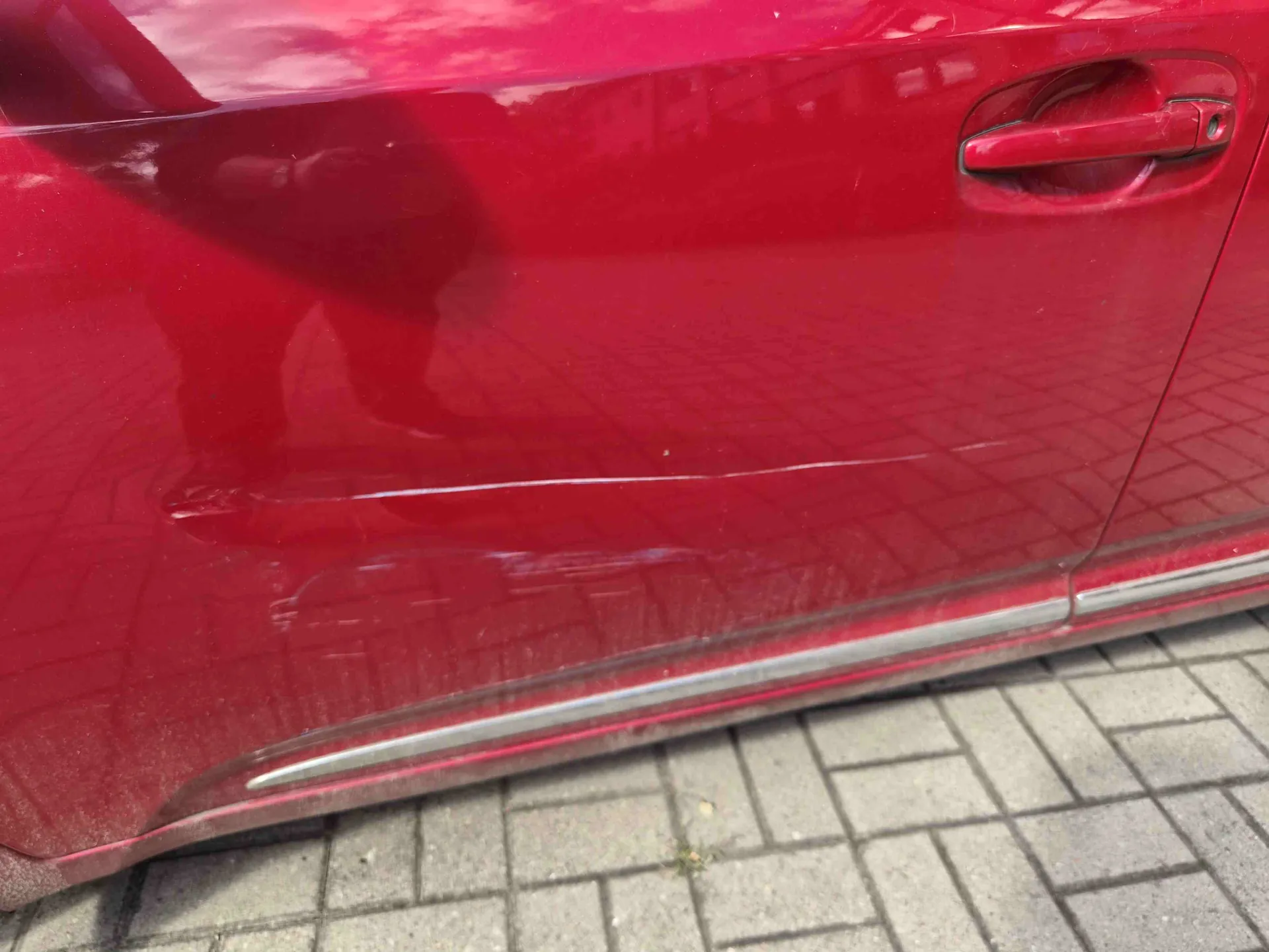 Zbliżenie na czerwone drzwi samochodu z widocznym wgnieceniem i zarysowaniem wzdłuż dolnej części panelu. Na lakierze widoczne są odbicia otoczenia oraz klamka zewnętrzna w prawym górnym rogu. Samochód stoi na szarym chodniku z kostki brukowej.