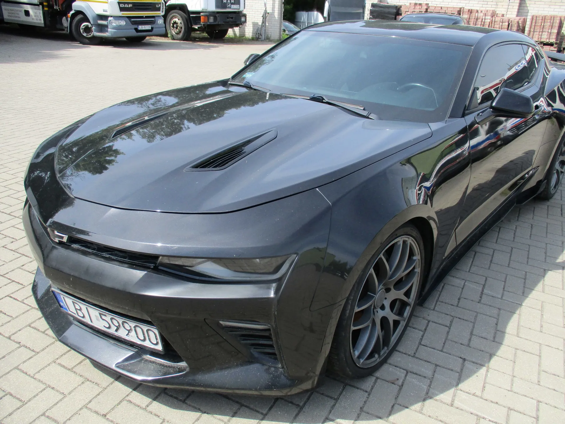 Czarny samochód sportowy Chevrolet Camaro zaparkowany na kostce brukowej. Widoczna jest przednia część pojazdu z tablicą rejestracyjną o numerze LBI 59900. W tle, w górnej części obrazu, widoczny jest fragment białej ciężarówki marki DAF.