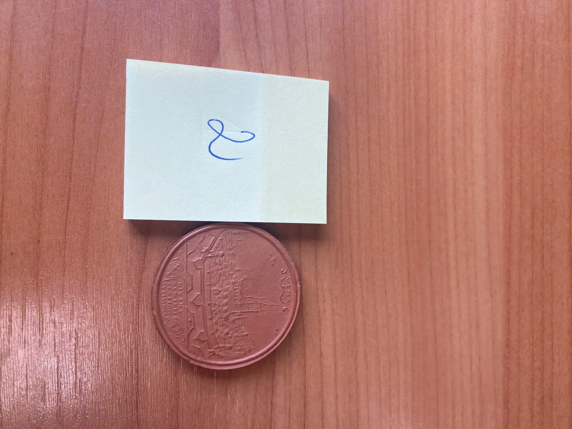 Na drewnianej powierzchni znajduje się jasnozielony karteczkowy notatnik z niebieskim symbolem ampersandu (&) napisanym na nim. Poniżej notatnika leży miedziana moneta z wygrawerowanym motywem budynku oraz widocznym rokiem "2016".