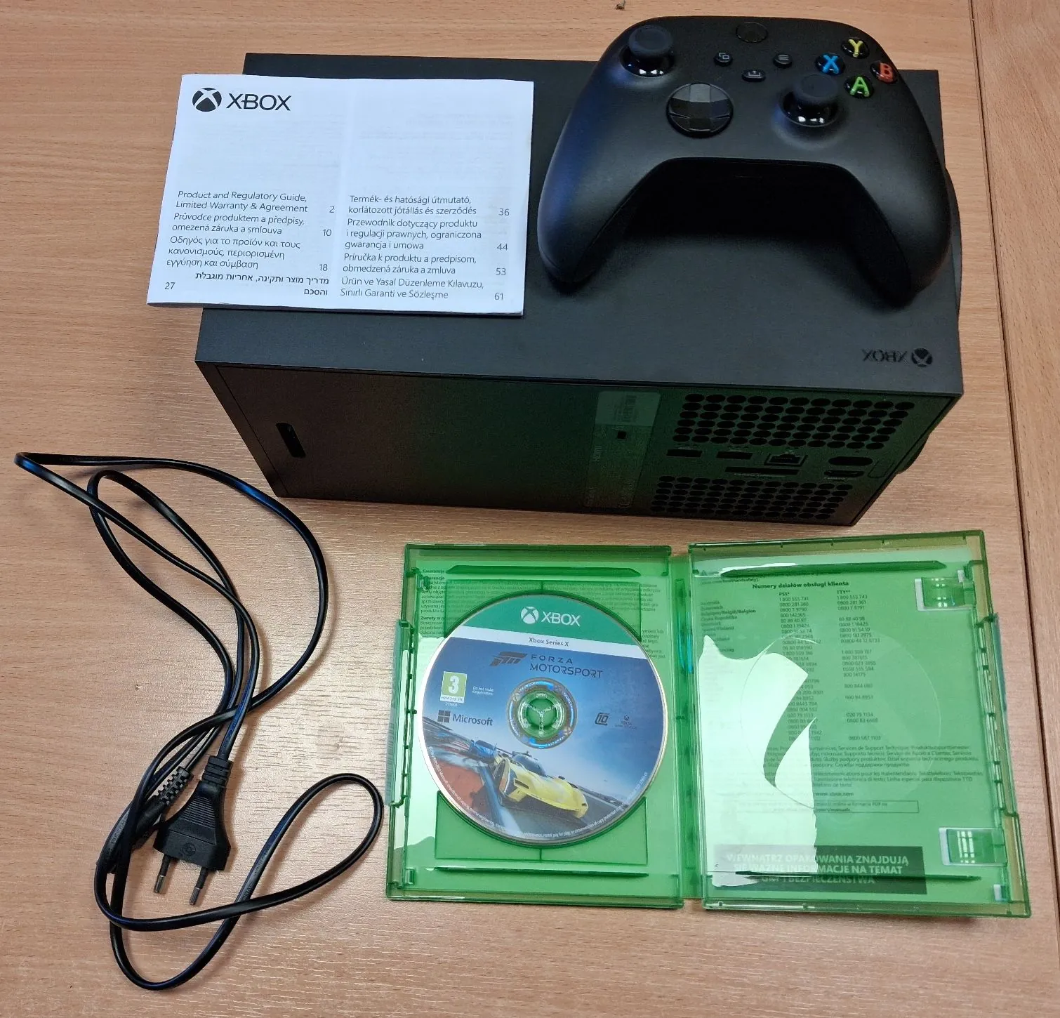 Czarna konsola Xbox Series S z czarnym kontrolerem leżącym na górze. Obok konsoli znajduje się otwarta plastikowa koperta z płytą do gry 'Forza Motorsport' oraz instrukcją obsługi. Przed konsolą leży czarny kabel zasilający. W lewym górnym rogu konsoli widoczna jest biała broszura z tekstem w wielu językach i logo Xbox.