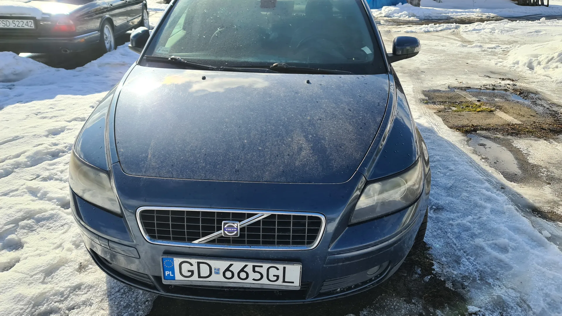 Ciemnoniebieski samochód marki Volvo z przodu, z tablicą rejestracyjną GD 665GL (PL) na zimowym parkingu. Na tle widoczny czarny samochód z tablicą SD 52242. Podłoże pokryte śniegiem i lodem z odsłoniętymi fragmentami asfaltu. Na masce samochodu widoczne ślady kurzu lub śniegu. Logo Volvo na atrapie chłodnicy.