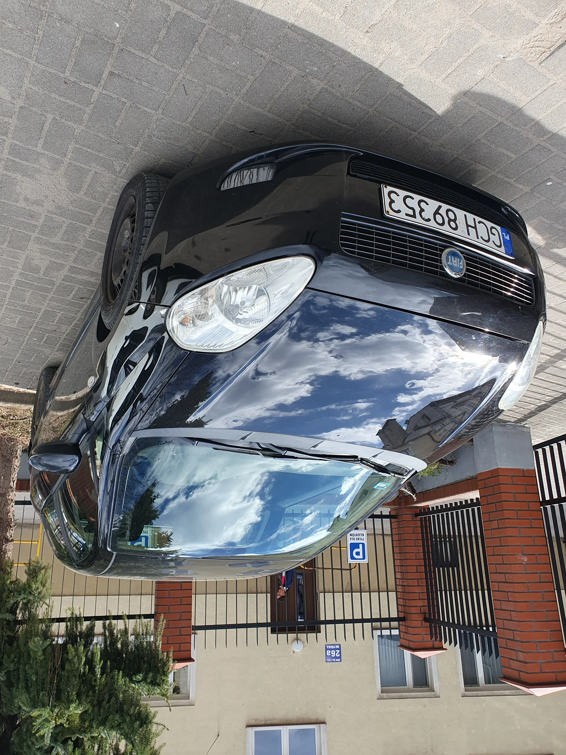 Czarny samochód marki Fiat zaparkowany na brukowanej powierzchni. Widoczna tablica rejestracyjna GCH 89353. W tle budynek z beżową elewacją, ceglanymi filarami i czarnym metalowym ogrodzeniem. Na budynku niebieski znak z napisem 26a oraz biało-niebieski znak parkingowy z literą P. Na przedniej szybie odbicie nieba z chmurami. Po lewej stronie drzewo iglaste.