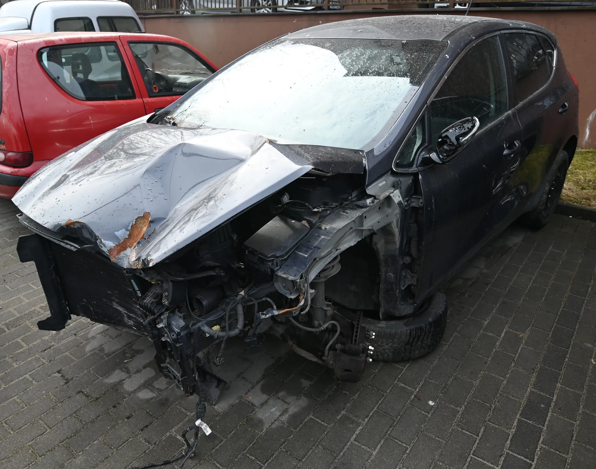 Zniszczony czarny samochód osobowy z silnie deformowaną przodem, pękniętym przednim szybą i odsłoniętym silnikiem. Widoczne uszkodzenia obejmują zgniecioną maskę z plamami rdzy, zniszczony zderzak oraz odsłonięte przewody. W tle czerwony samochód i biały van zaparkowane przy brązowej ścianie. Samochody stoją na brukowanej nawierzchni, po prawej stronie fragment trawnika.