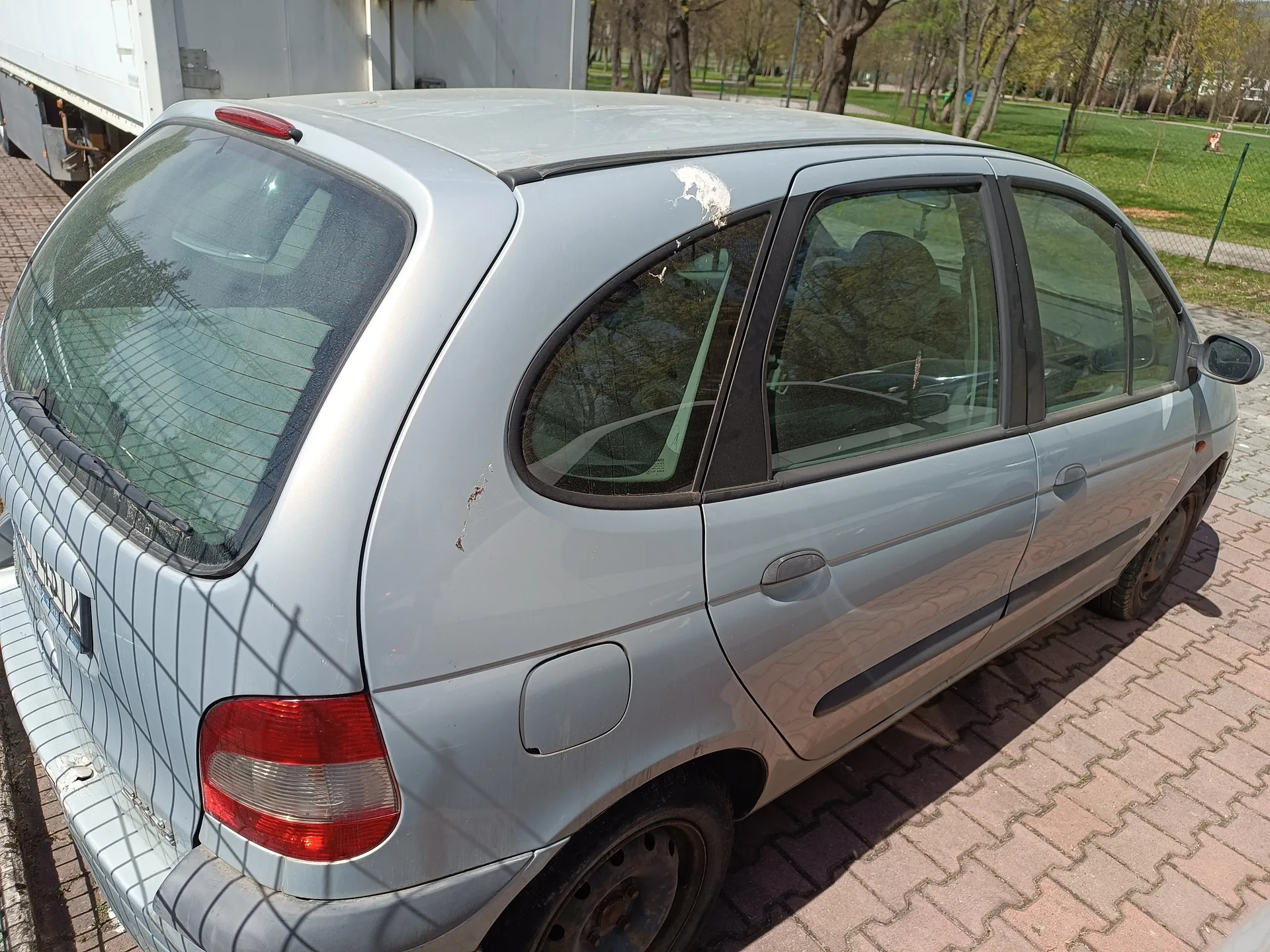 Srebrny samochód osobowy typu hatchback zaparkowany na brukowanej powierzchni. Widoczne uszkodzenia na dachu i tylnej części nadwozia, w tym zarysowana farba. Tylne światło czerwone, tablica rejestracyjna częściowo widoczna. W tle biały naczepowy pojazd ciężarowy, drzewa, trawnik, ogrodzenie i chodnik. Słońce rzuca cienie na nadwozie i bruk.
