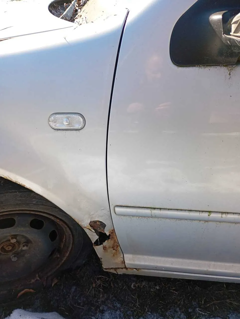 Boczny widok białego samochodu z widocznym uszkodzeniem i rdzą w okolicy przedniego koła. Widoczne są zardzewiały felga, czarna obudowa lusterka bocznego oraz zielone naloty na karoserii. Pod samochodem znajduje się trawa i resztki śniegu.