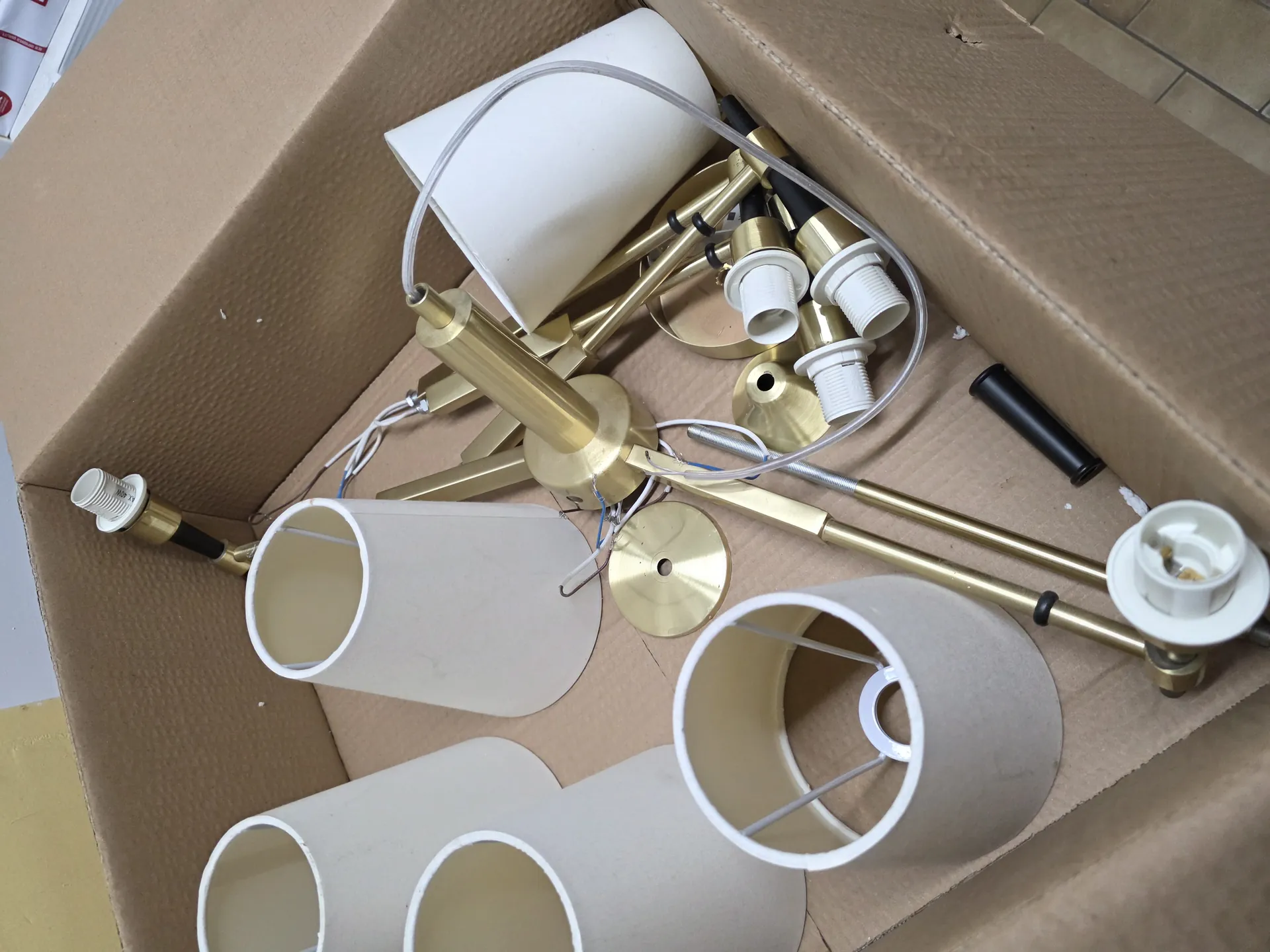 Otwarte kartonowe pudełko zawierające elementy lampy: mosiężne metalowe części, białe cylindryczne abażury, przewody elektryczne, czarne i białe gniazda oraz inne małe komponenty. Elementy są ułożone wewnątrz pudełka bez określonego porządku.